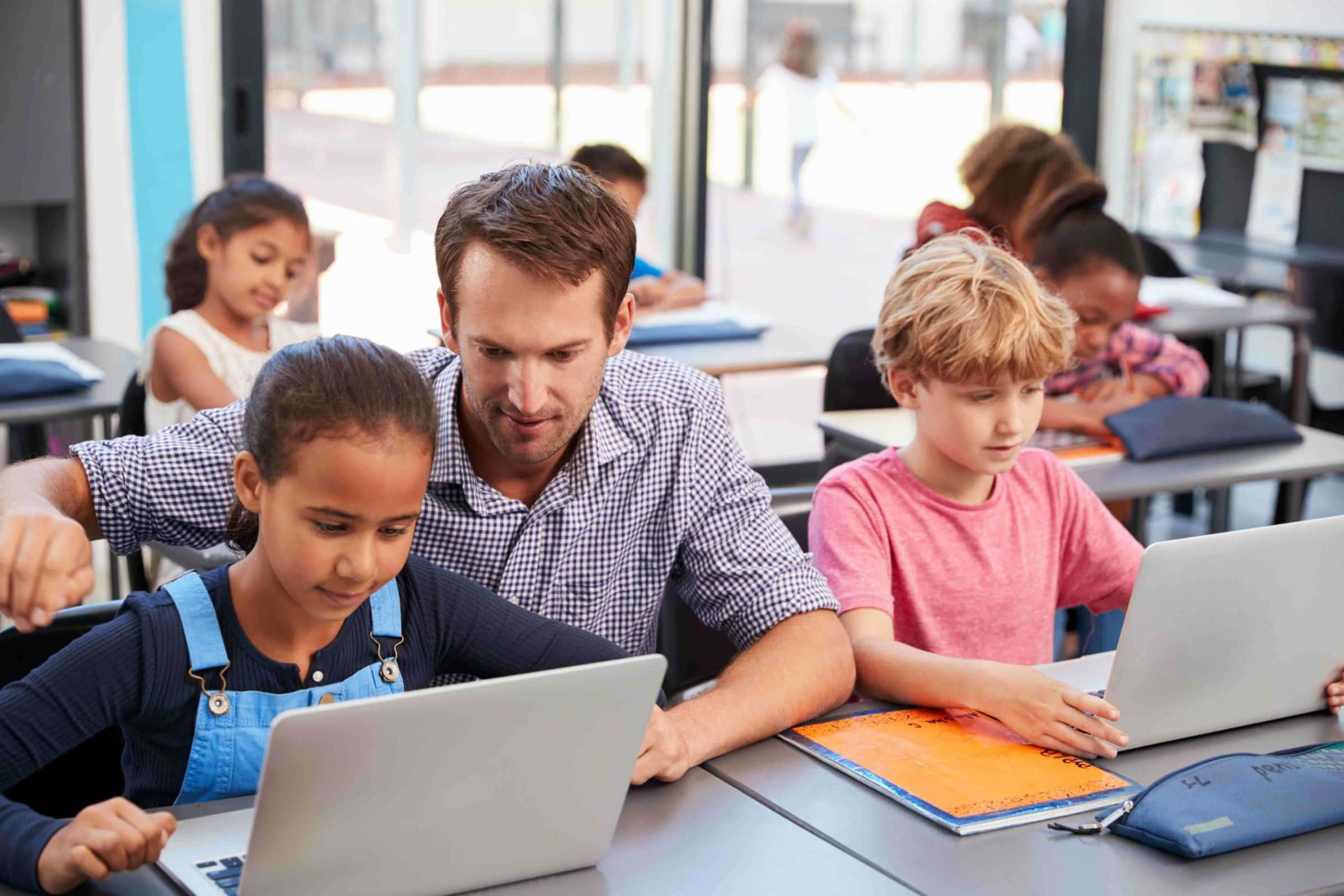 Classroom 桌布 壁紙 — 一位老師俯身協助一位正在使用筆記型電腦的小女孩，教室明亮現代，其他學生專注於自己的書桌上。