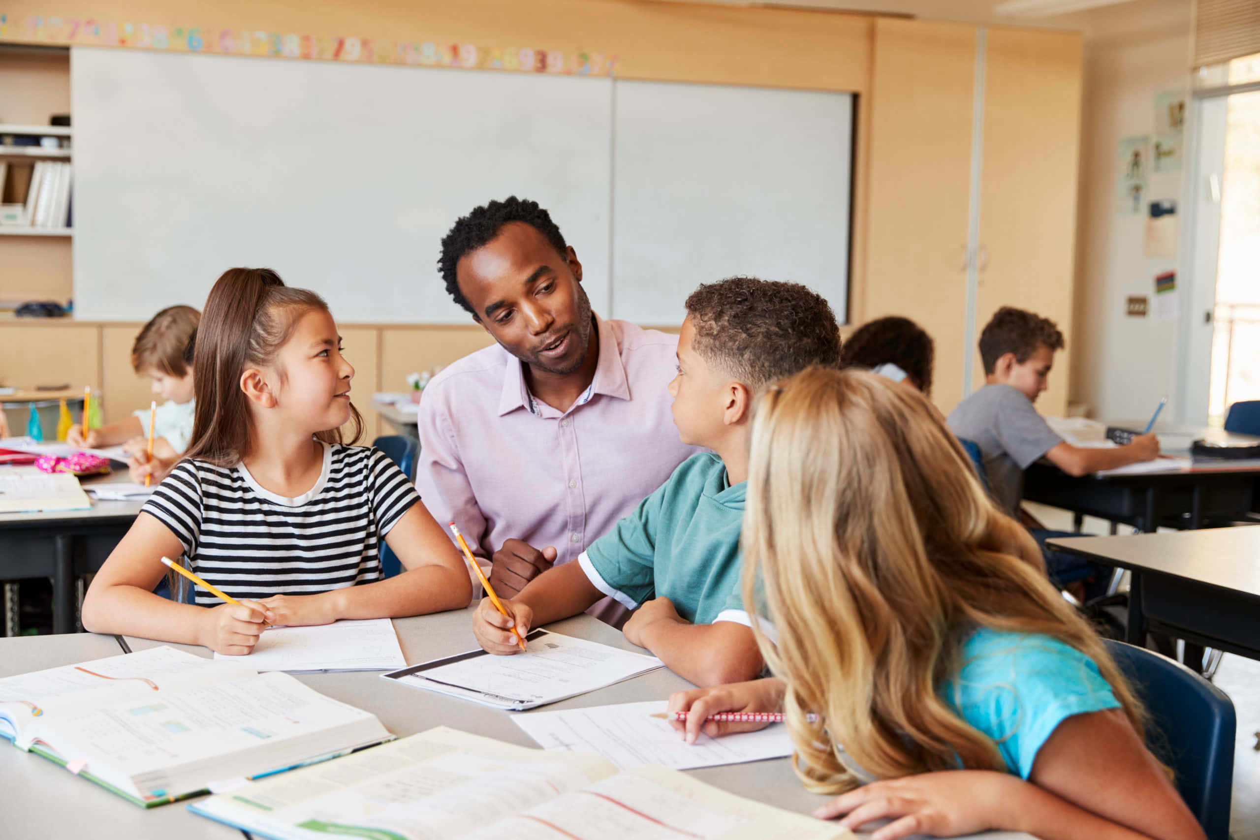 Classroom 桌布 壁紙 — 一位老師俯身幫助坐在書桌前的學生，明亮的教室裡放滿了打開的書本和鉛筆。