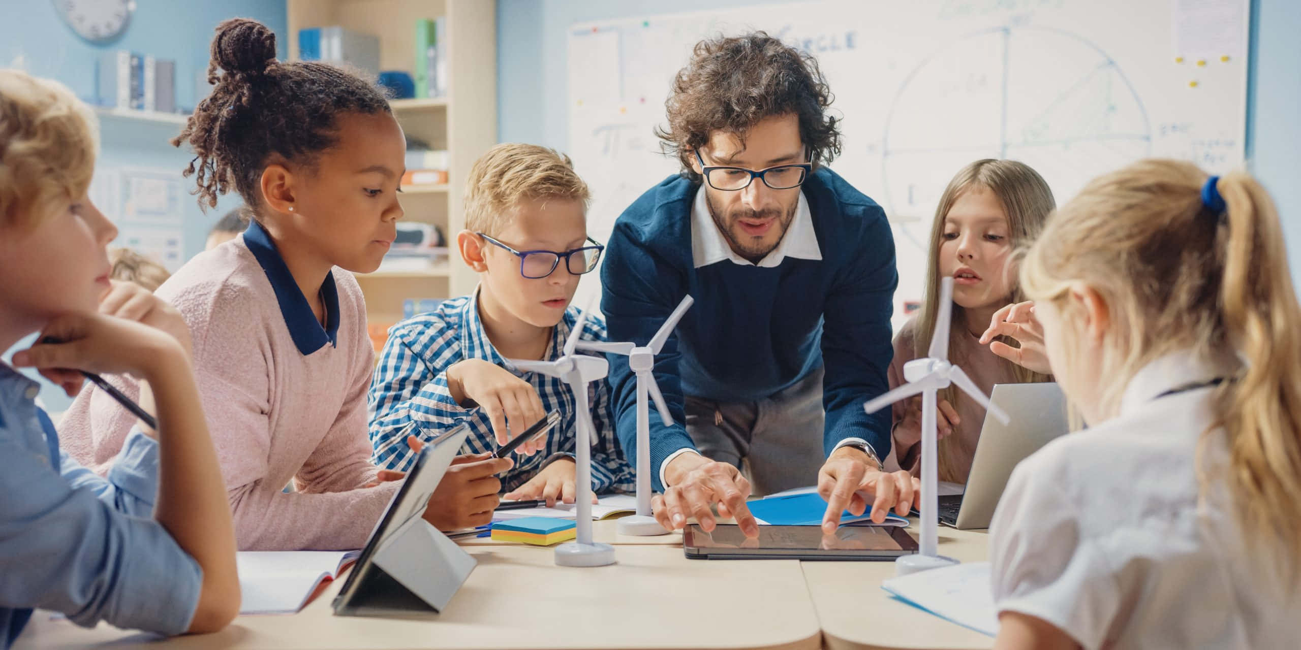 Classroom 桌布 壁紙 — 一位老師在明亮的教室內，引導不同學生圍繞一張放有風力渦輪機模型和平板電腦的桌子。