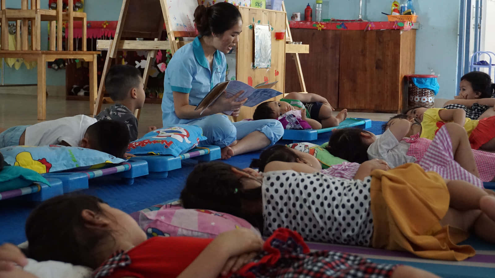 People Sleeping 桌布 壁紙 — 一位老師在明亮的教室裡，為躺在彩色地墊上休息的孩子們讀故事。