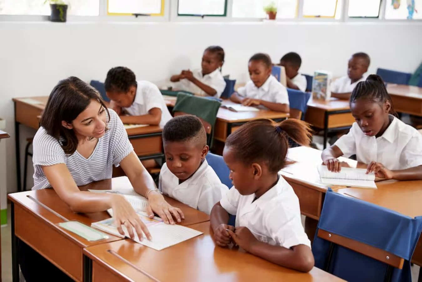 Education 桌布 壁紙 — 一位老師在明亮的教室裡俯身靠近學生的書桌，其他孩子則專注於自己的作業。