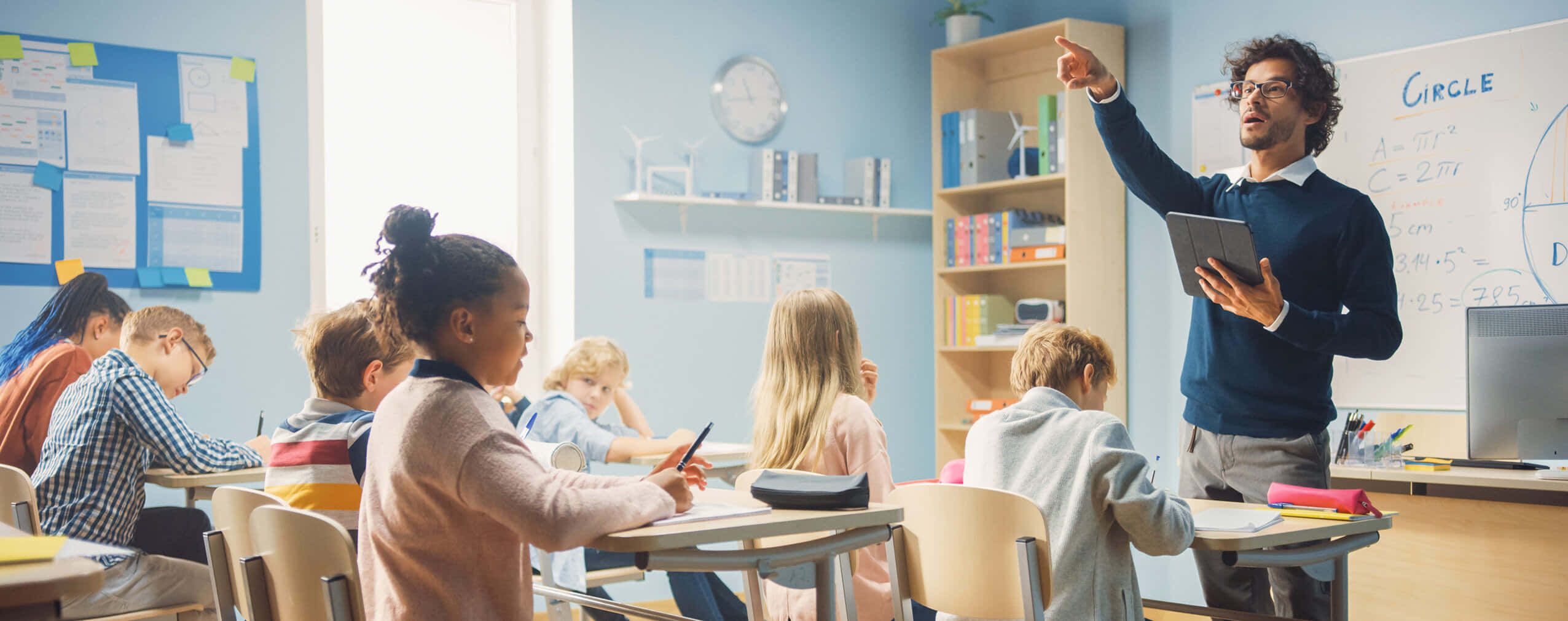 Classroom 桌布 壁紙 — 一位老師指著白板，學生們坐在書桌前，教室明亮，充滿教育海報和書籍。