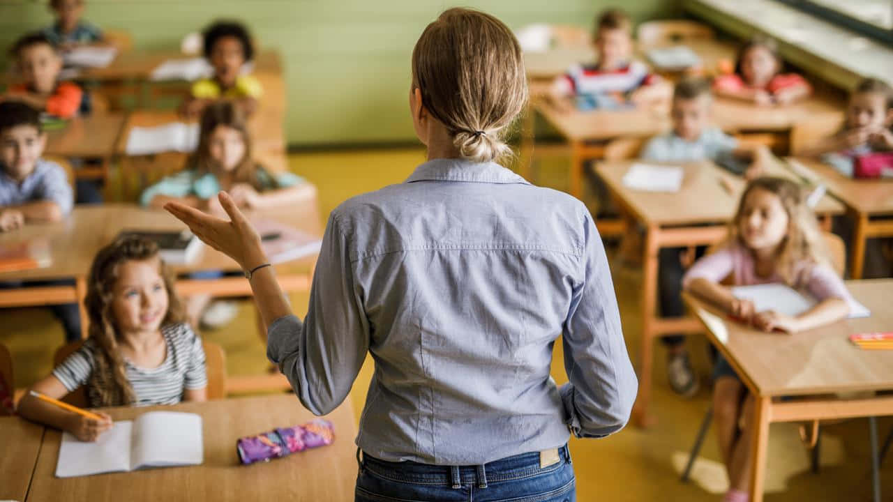 Learning 桌布 壁紙 — 一位老師背對鏡頭站著，向坐在明亮教室裡木桌旁的學生做手勢。