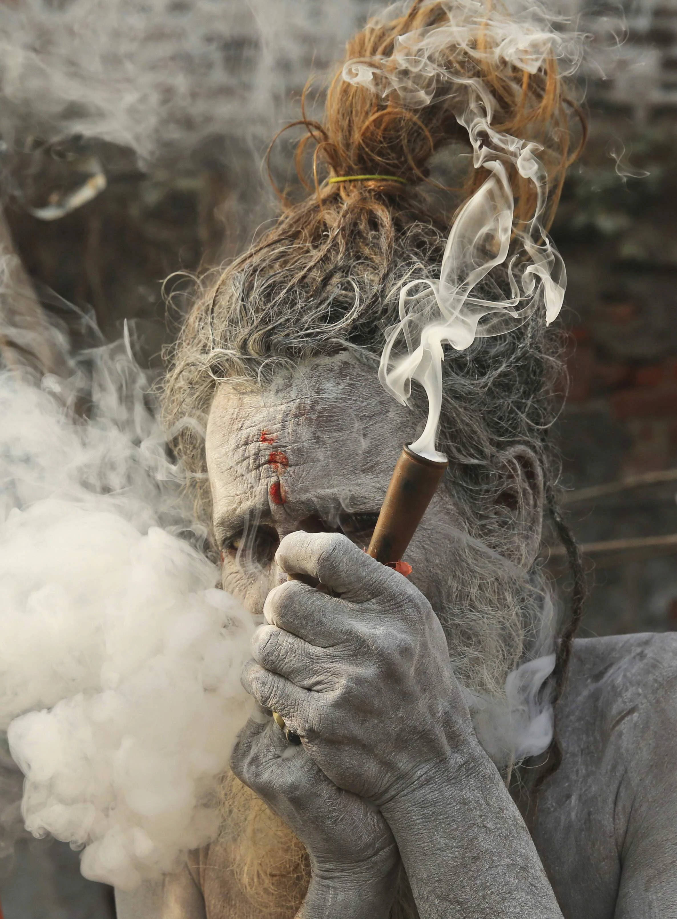 Aghori 桌布 壁紙 — 一位聖人，皮膚覆蓋灰燼，頭髮凌亂，對著模糊的磚牆從煙斗中呼出濃煙。