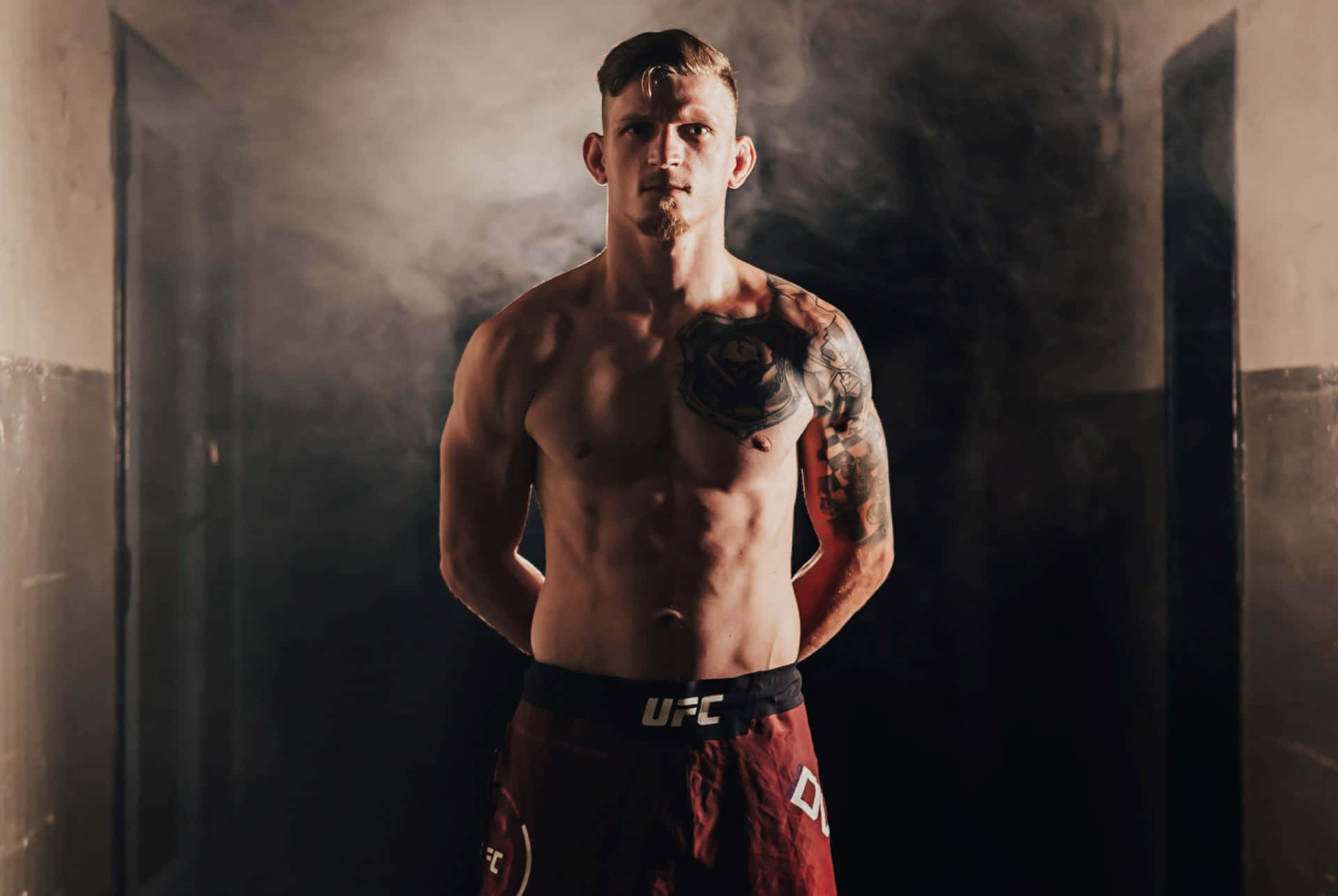 UFC 桌布 壁紙 — 一位肌肉發達、帶有刺青的UFC拳手，自信地站在煙霧瀰漫、燈光昏暗的走廊中，穿著紅色格鬥短褲。