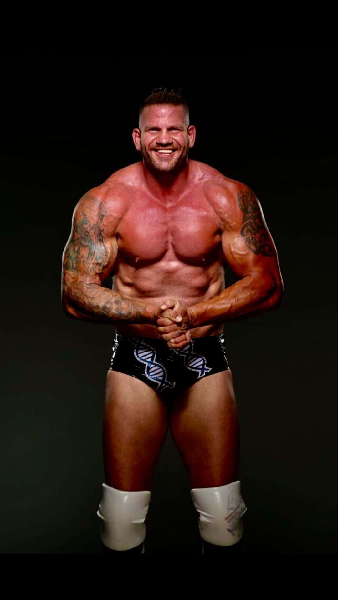 WWE 桌布 壁紙 — 一位肌肉發達、帶有紋身的摔角手，穿著黑色三角褲和白色護膝，在黑暗的攝影棚背景下自信地微笑。