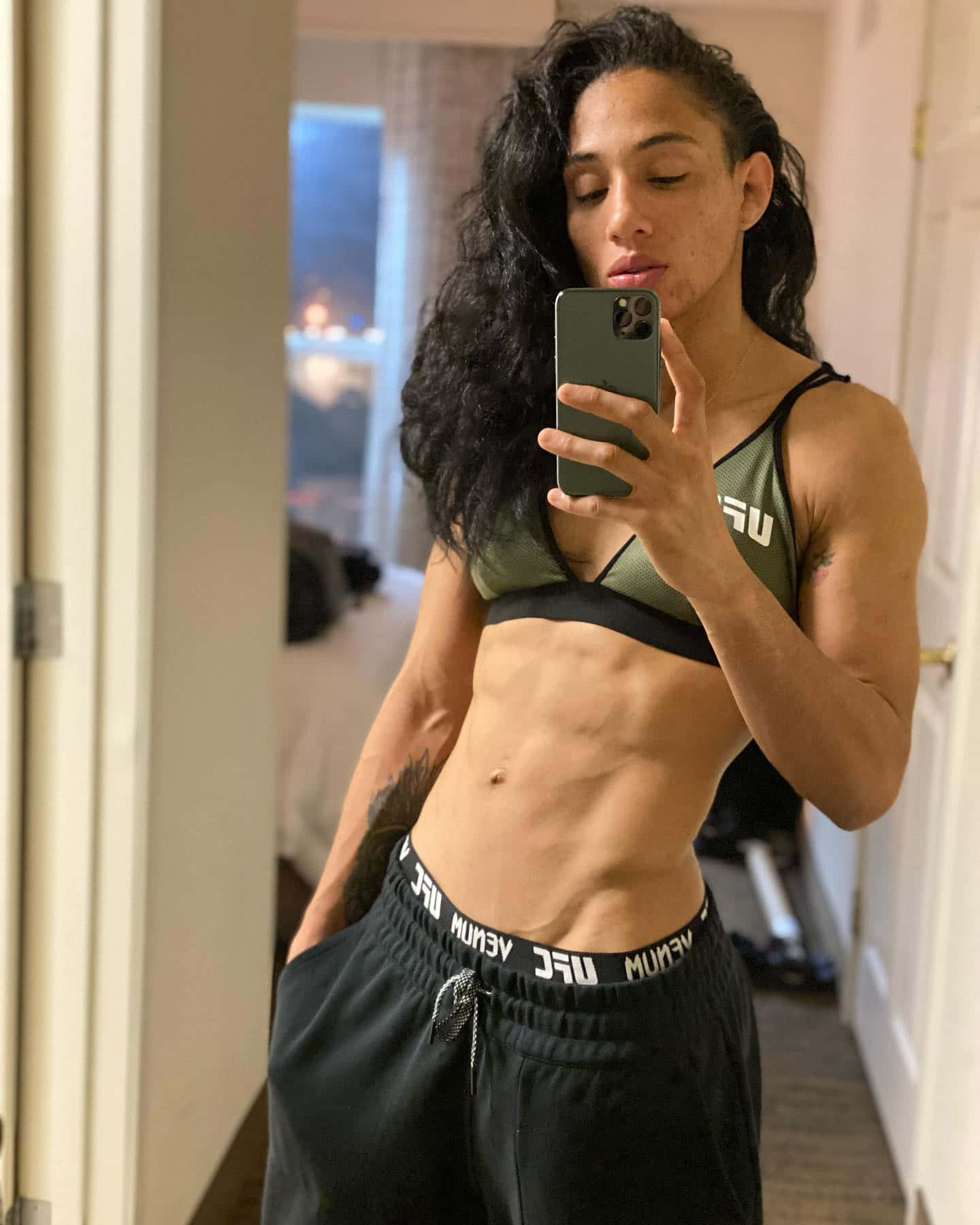 MMA 桌布 壁紙 — 一位肌肉發達、捲髮的女性穿著UFC裝備，在昏暗的走廊裡對著鏡子自拍，展示她線條分明的腹肌。