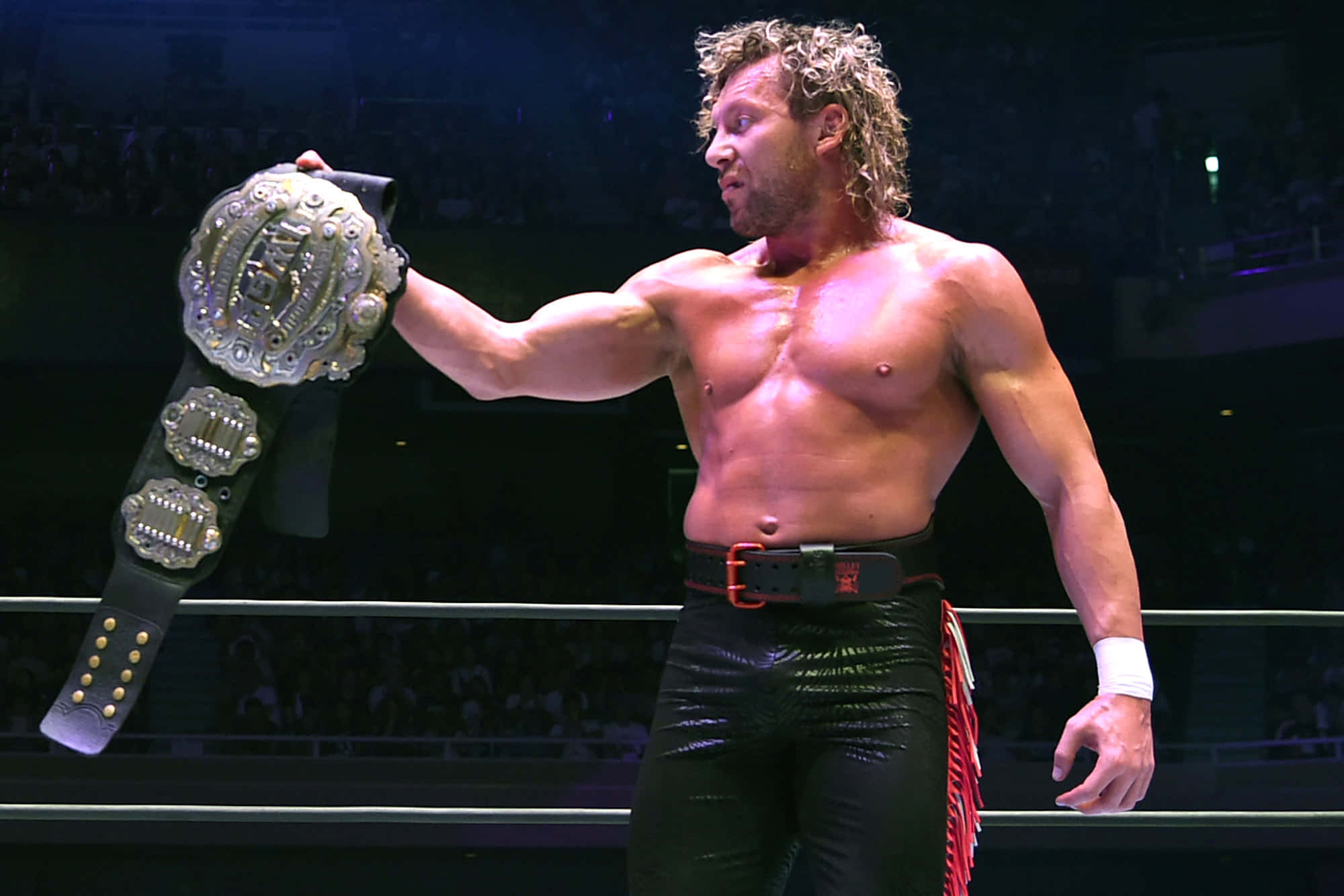 Aew 桌布 壁紙 — 一位肌肉發達、捲髮的摔角手在燈光明亮的擂台上自豪地高舉冠軍腰帶，身後是歡呼的人群。