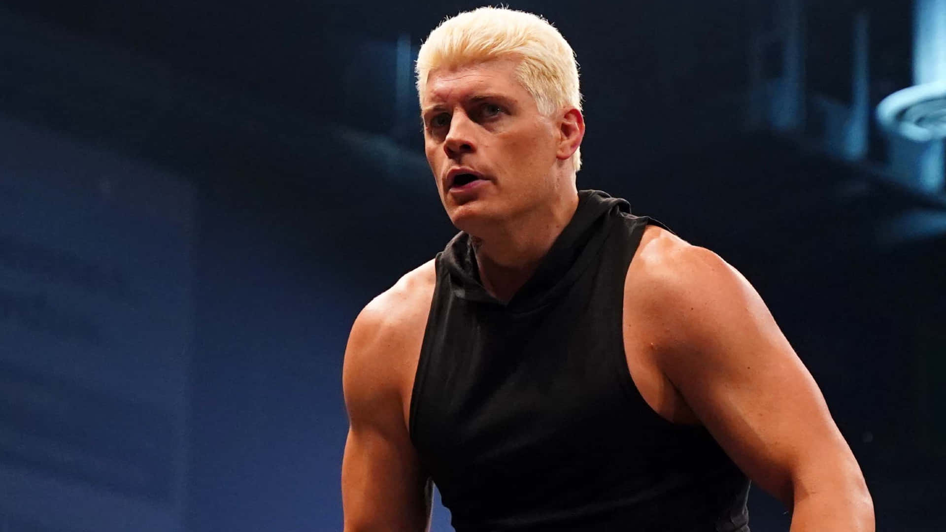 WWE 桌布 壁紙 — 一位肌肉發達、擁有鉑金色頭髮且表情強烈的男子，在戲劇性的舞台燈光下穿著黑色背心。