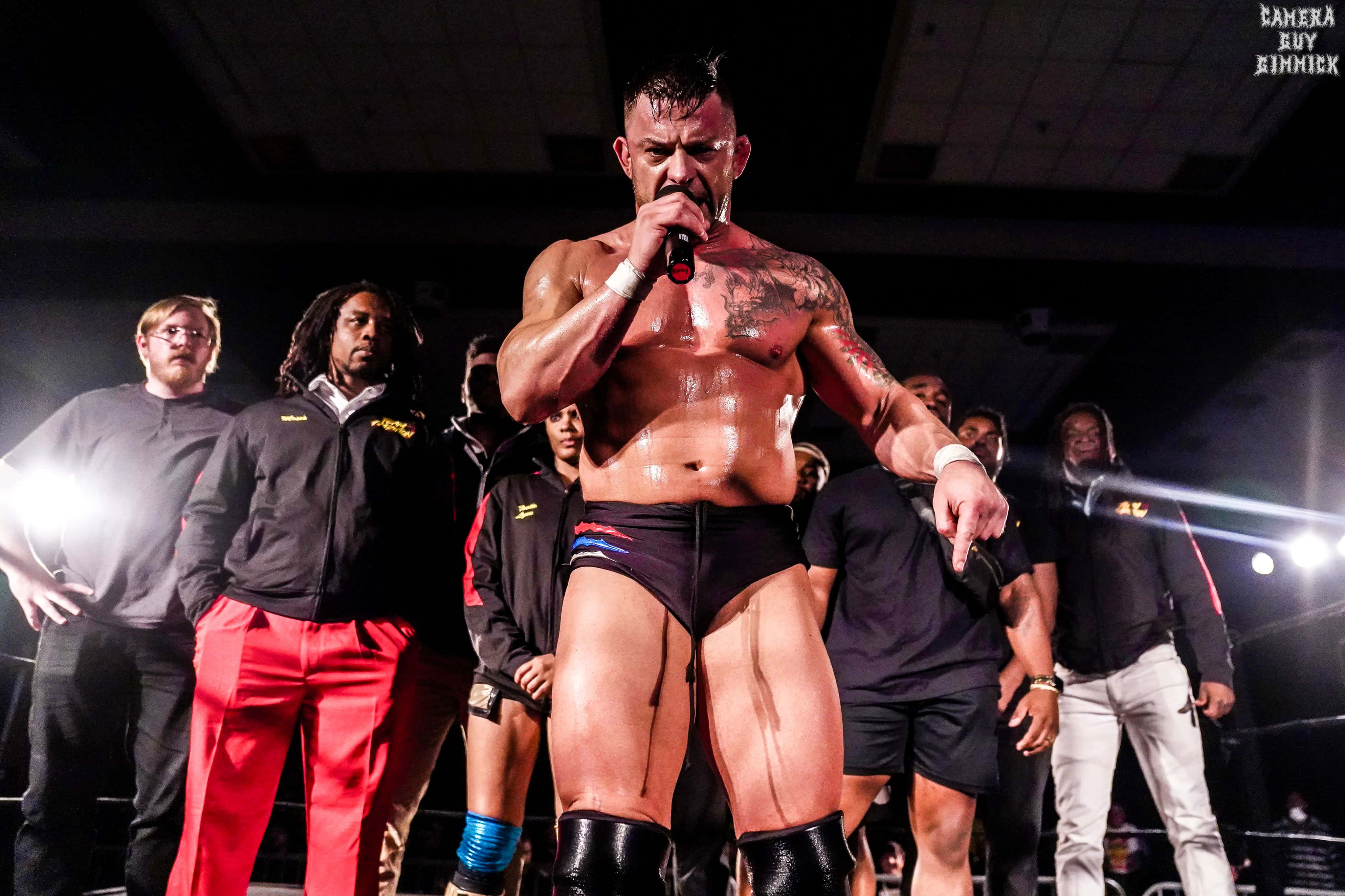 WWE 桌布 壁紙 — 一位肌肉發達、有刺青、穿黑色靴子的摔角手，在明亮的舞台燈光下拿著麥克風，激烈地指向人群。