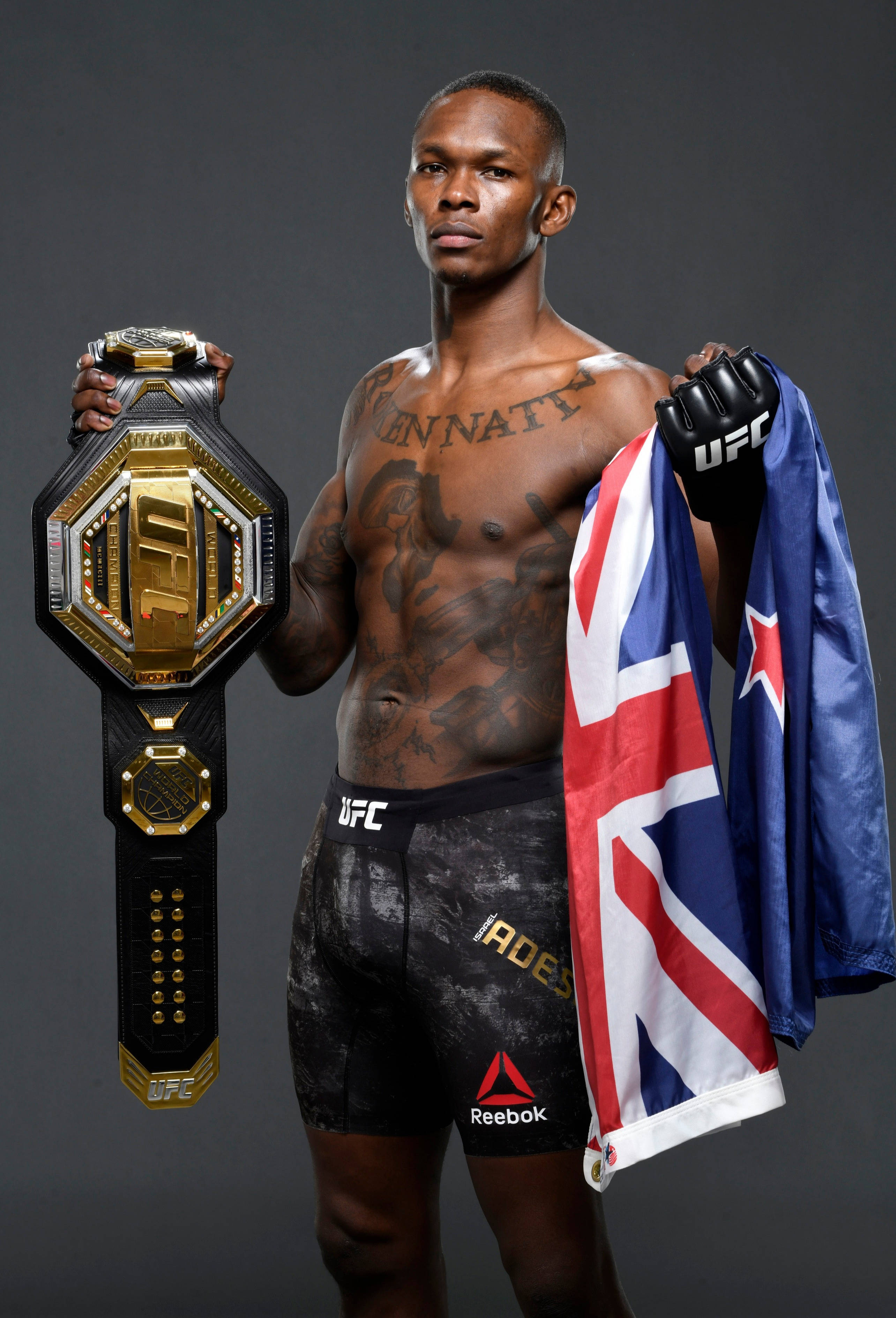 Adesanya 桌布 壁紙 — 一位肌肉發達、有刺青的UFC拳手在黑暗的攝影棚背景下，手持冠軍腰帶和英國國旗。