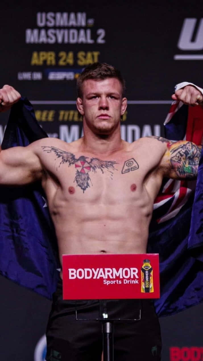 UFC 桌布 壁紙 — 一位肌肉發達、有刺青的拳手在UFC稱重活動中赤裸上身站在體重計上，手持旗幟。