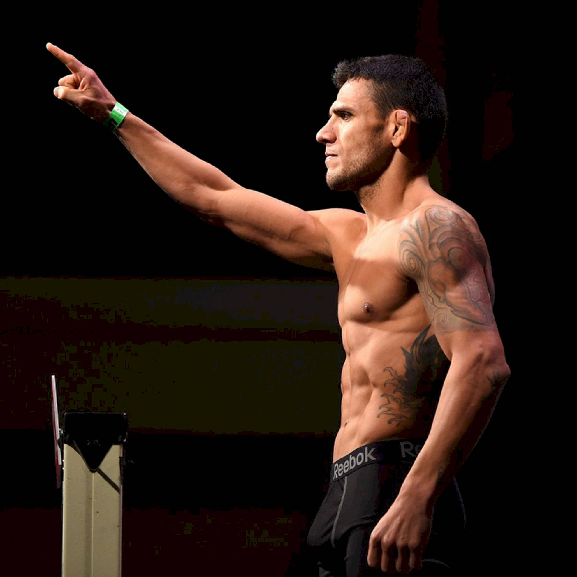 MMA 桌布 壁紙 — 一位肌肉發達、有刺青的拳手在燈光昏暗的競技場上穿著Reebok短褲向前指。