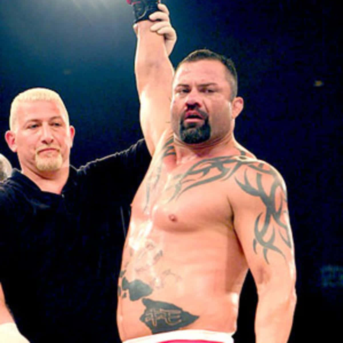 UFC 桌布 壁紙 — 一位肌肉發達、有刺青的摔角手在昏暗的競技場中勝利地舉起手臂，裁判站在他旁邊。