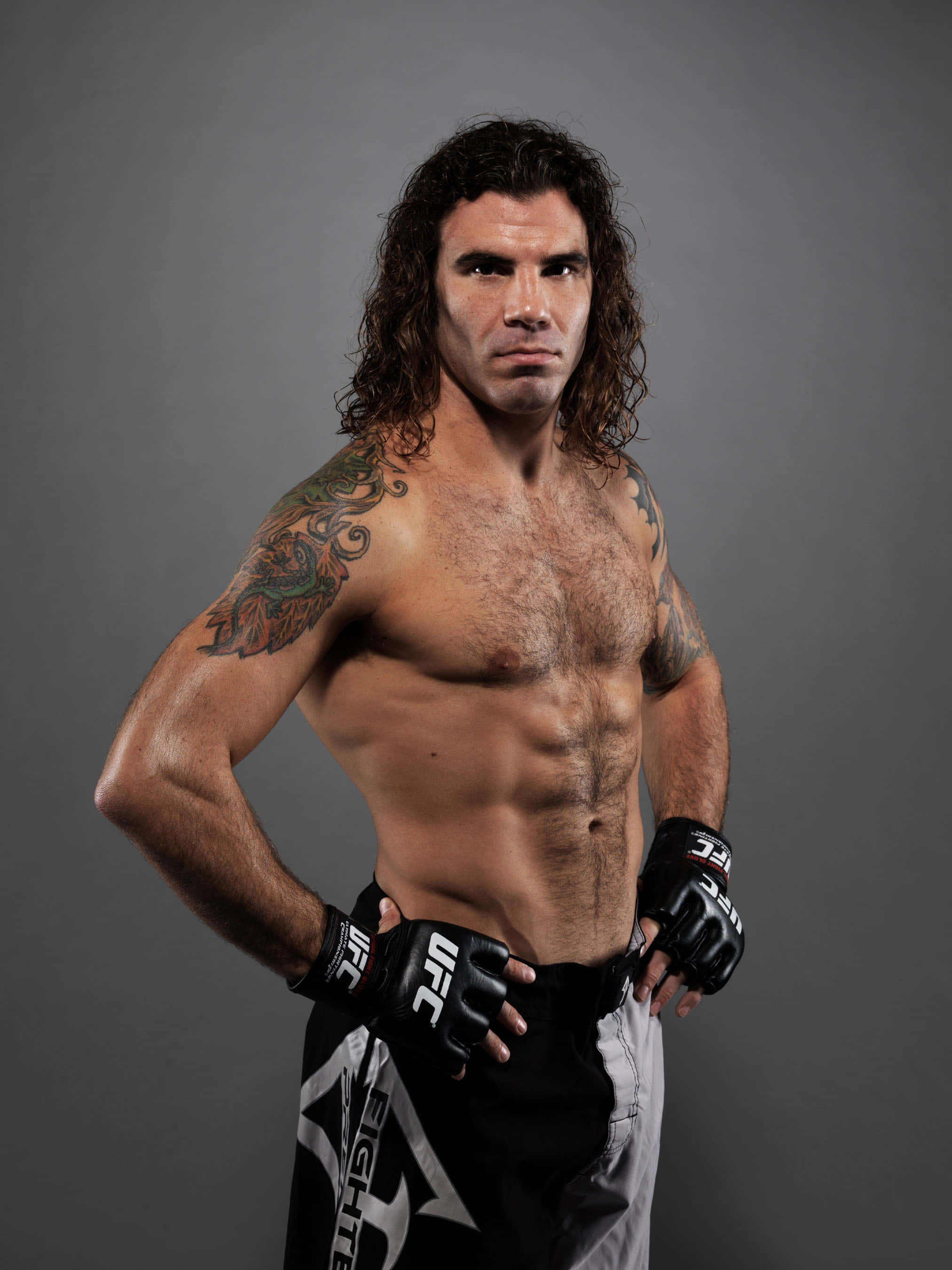 UFC 桌布 壁紙 — 一位肌肉發達、留著深色長髮和紋身的戰士，在灰色工作室背景下，戴著UFC手套和格鬥短褲，凝視著鏡頭。