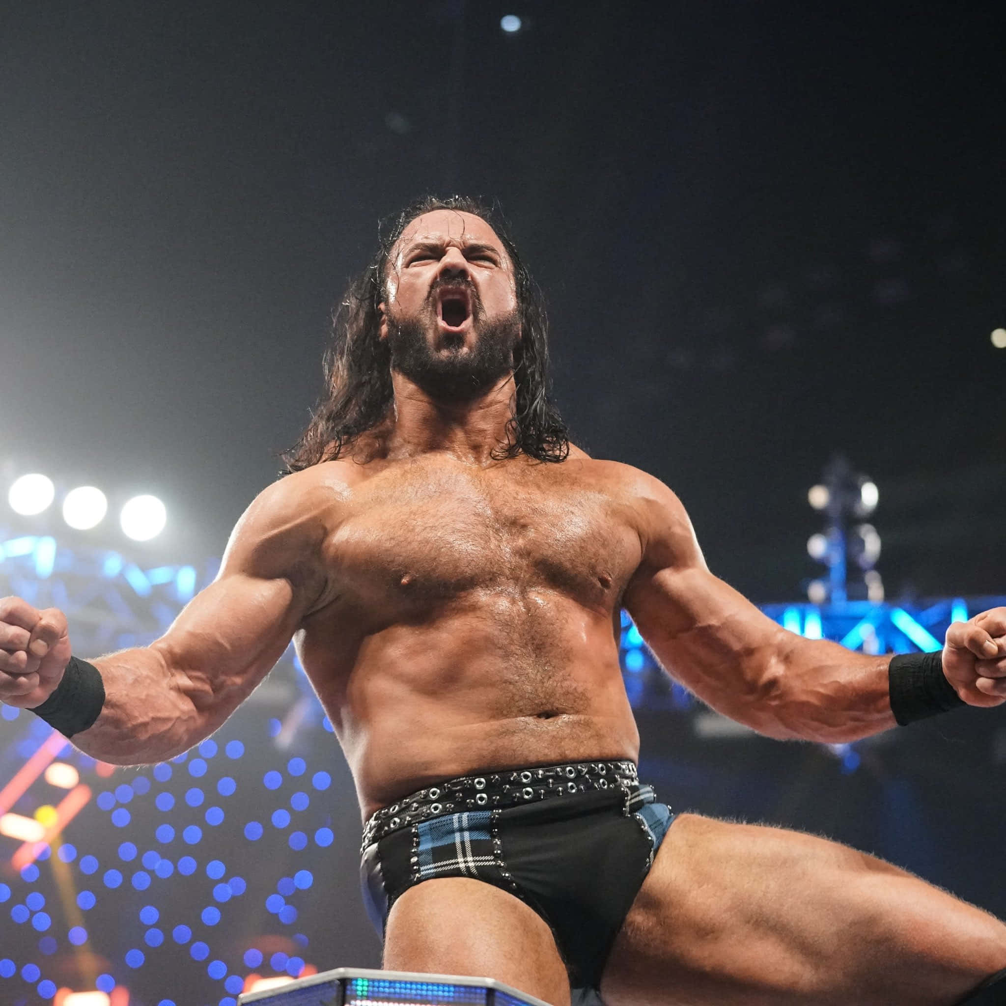 WWE 桌布 壁紙 — 一位肌肉發達、留著深色長髮和鬍子的摔角手，在身後藍色燈光的明亮擂台上勝利地咆哮。