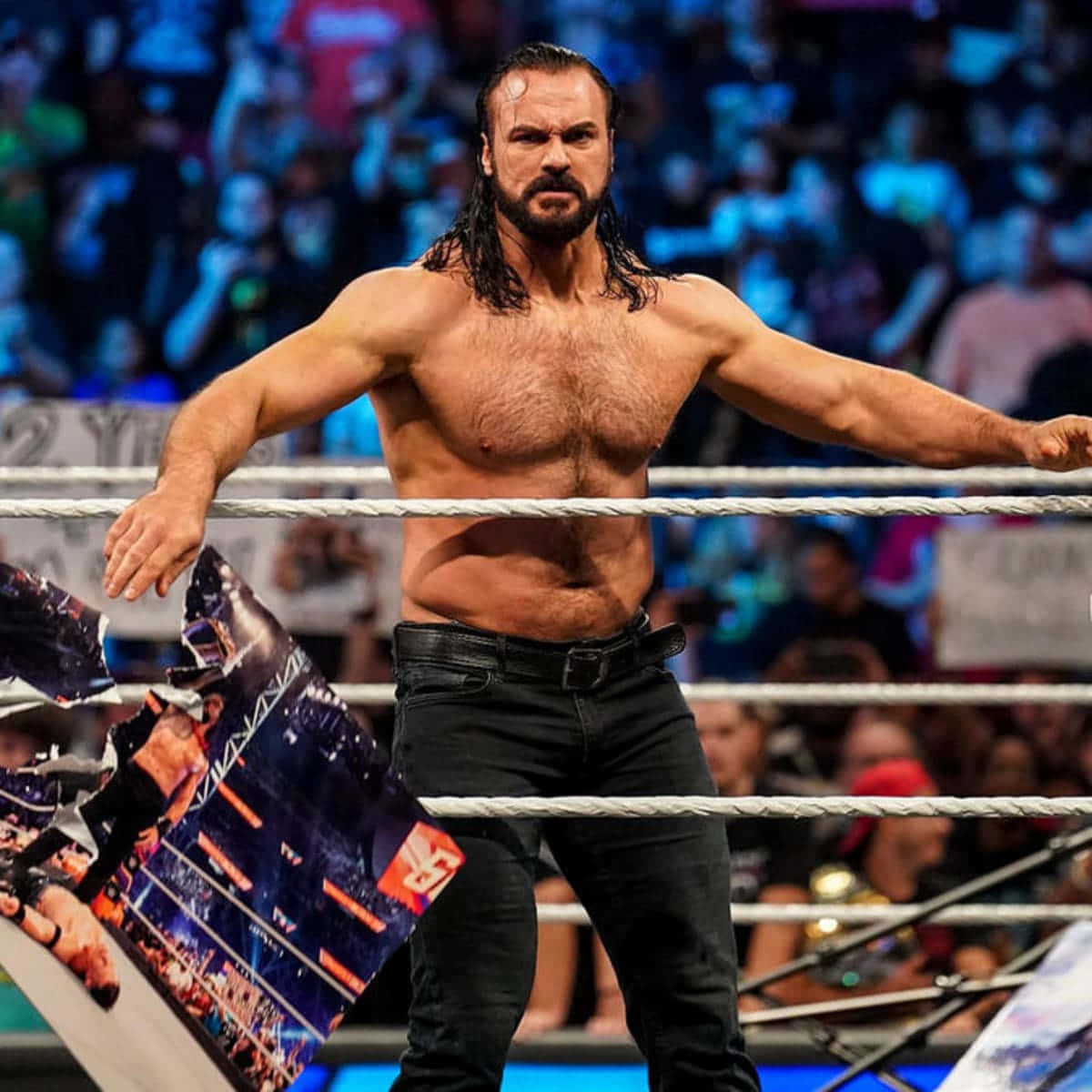 WWE 桌布 壁紙 — 一位肌肉發達、留著深色長髮和鬍鬚的摔角手站在摔角場上，手持撕破的海報，表情強烈。