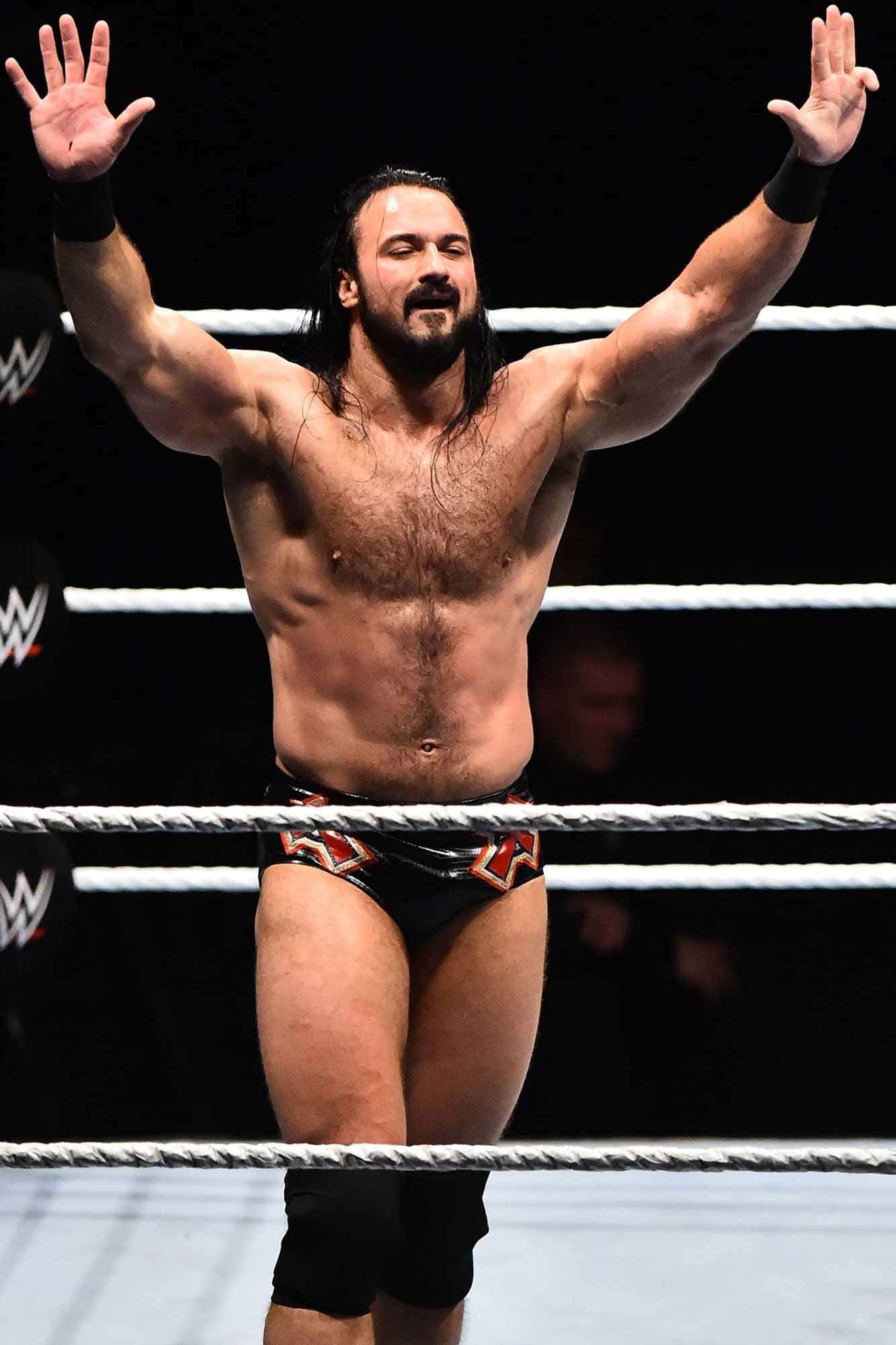 WWE 桌布 壁紙 — 一位肌肉發達、留著深色長髮和鬍鬚的摔角手，身穿帶有紅色和金色圖案的黑色短褲，在摔角場上高舉雙臂，展現勝利姿態。
