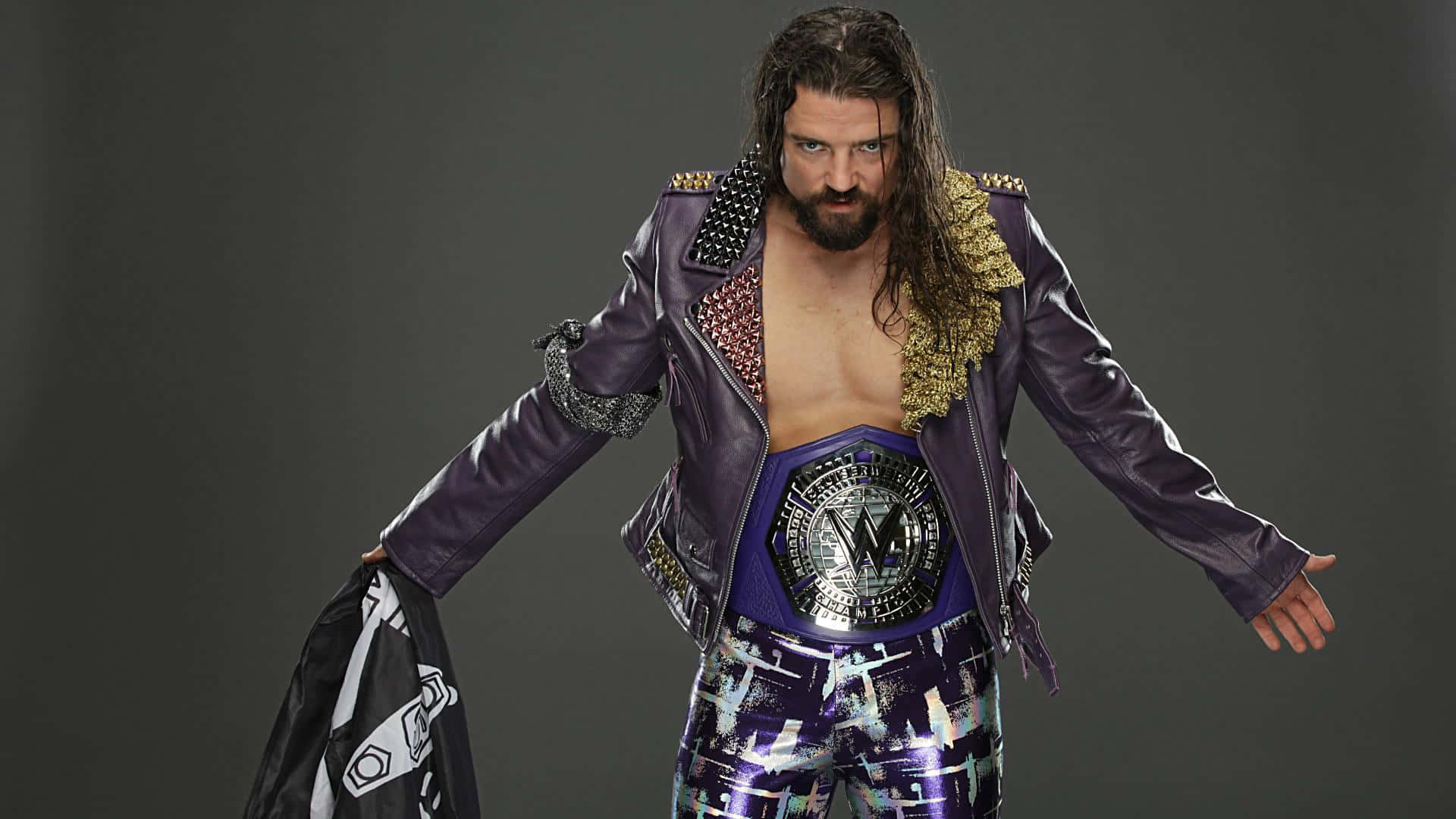 WWE 桌布 壁紙 — 一位肌肉發達、留著深色長髮和鬍鬚的摔角手，穿著紫色鉚釘外套和冠軍腰帶，站在灰色攝影棚背景前。