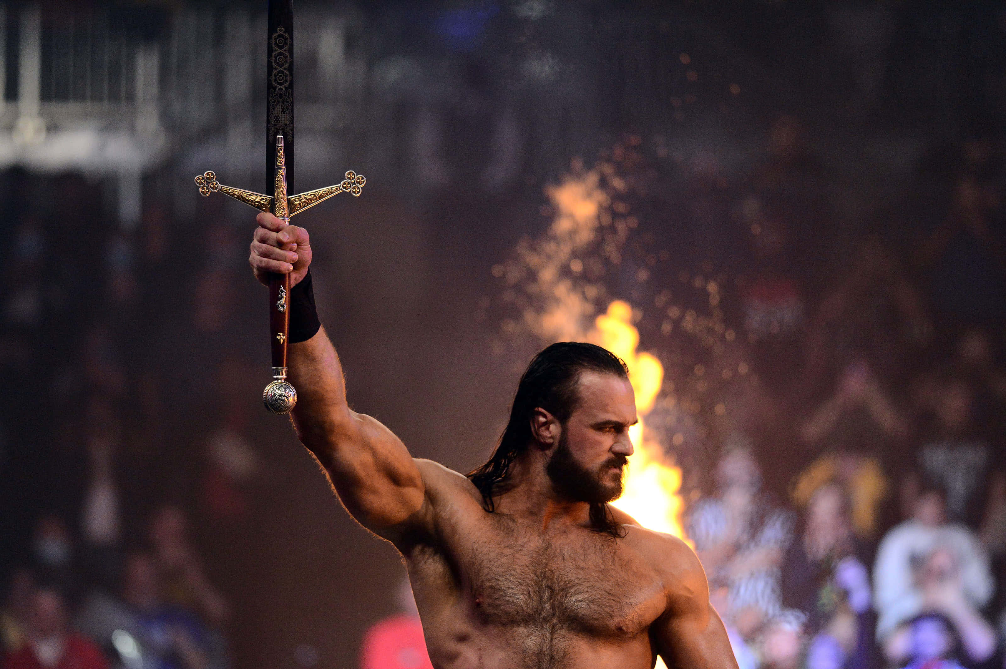 WWE 桌布 壁紙 — 一位肌肉發達、留著深色長髮的摔角手，在身後咆哮的人群和熾熱的煙火前，將一把華麗的金劍高舉過頭。