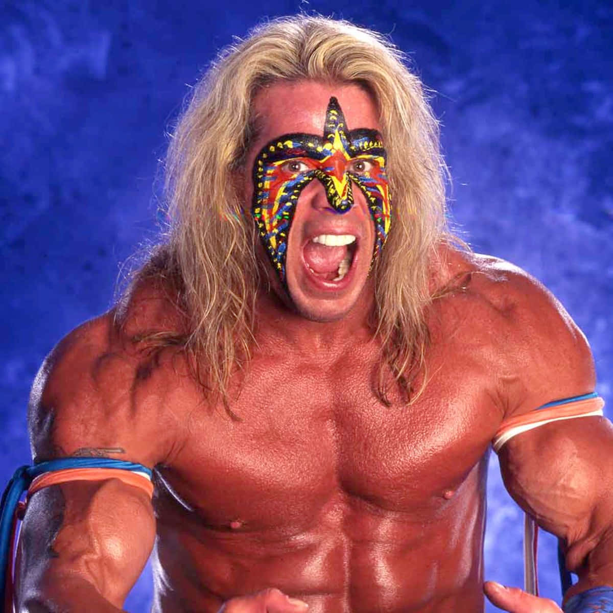 WWE 桌布 壁紙 — 一位肌肉發達、留著金色長髮、臉上畫著彩色星星圖案的摔角手，在深藍色背景下猛烈咆哮。