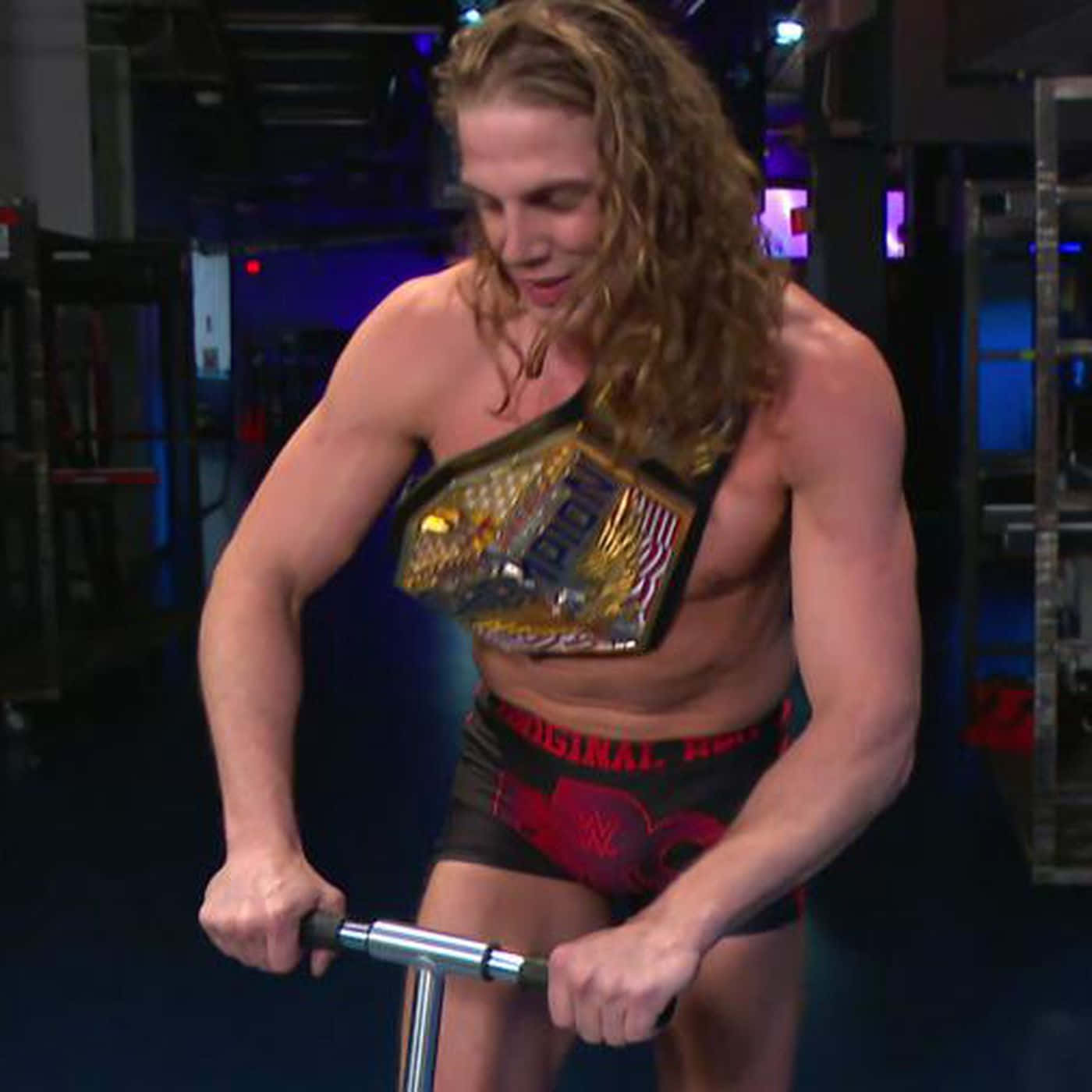 WWE 桌布 壁紙 — 一位肌肉發達、留著金色長髮的摔角手，在燈光昏暗的健身房裡，戴著冠軍腰帶騎固定腳踏車。