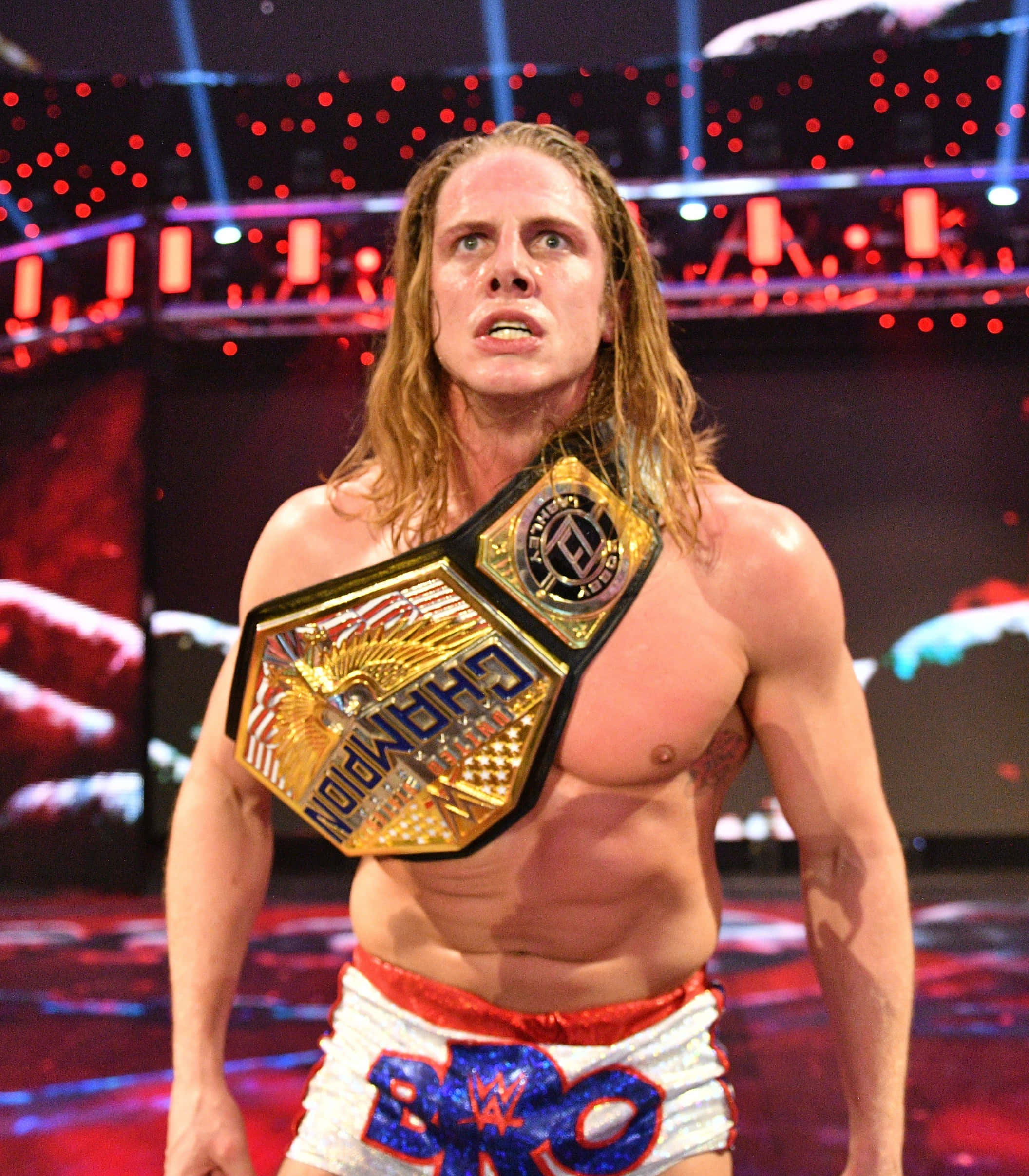 WWE 桌布 壁紙 — 一位肌肉發達、留著金色長髮的摔角手站在擂台上，戴著金色冠軍腰帶，穿著帶有藍色字母的白色短褲。