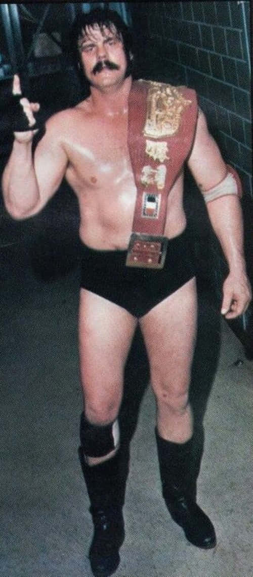 WWE 桌布 壁紙 — 一位肌肉發達、留著鬍子、腰間繫著冠軍腰帶的摔角手，在燈光昏暗的後台區域自信地擺姿勢。