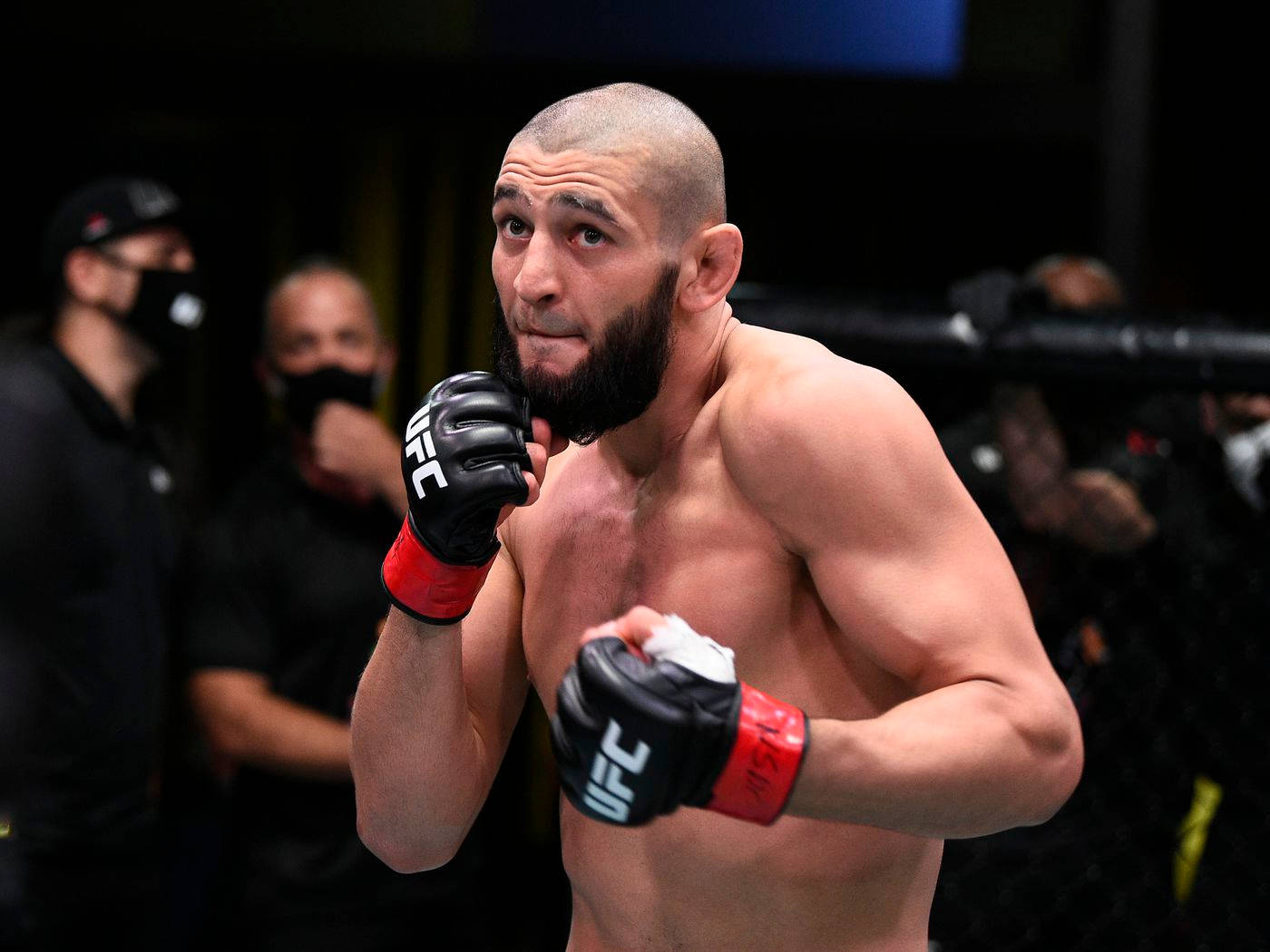 MMA 桌布 壁紙 — 一位肌肉發達、留鬍子的戰士戴著黑色和紅色的手套，在昏暗的八角籠中以專注的強度警覺地站立。
