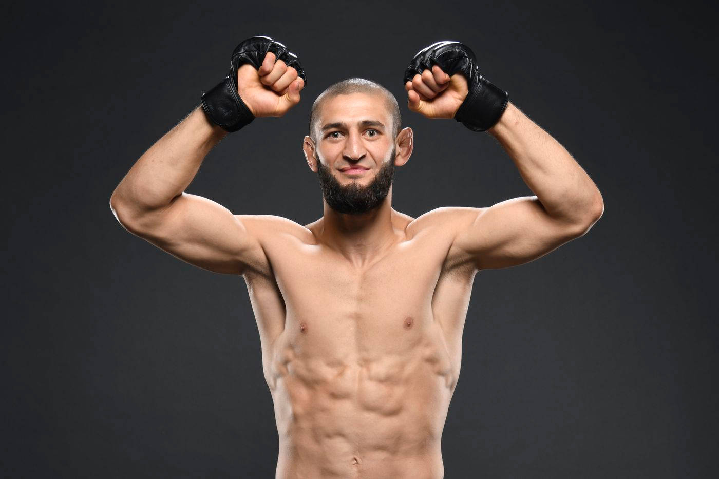 UFC 桌布 壁紙 — 一位肌肉發達、留鬍子的戰士戴著黑色手套，在黑暗的攝影棚背景下勝利地舉起拳頭。