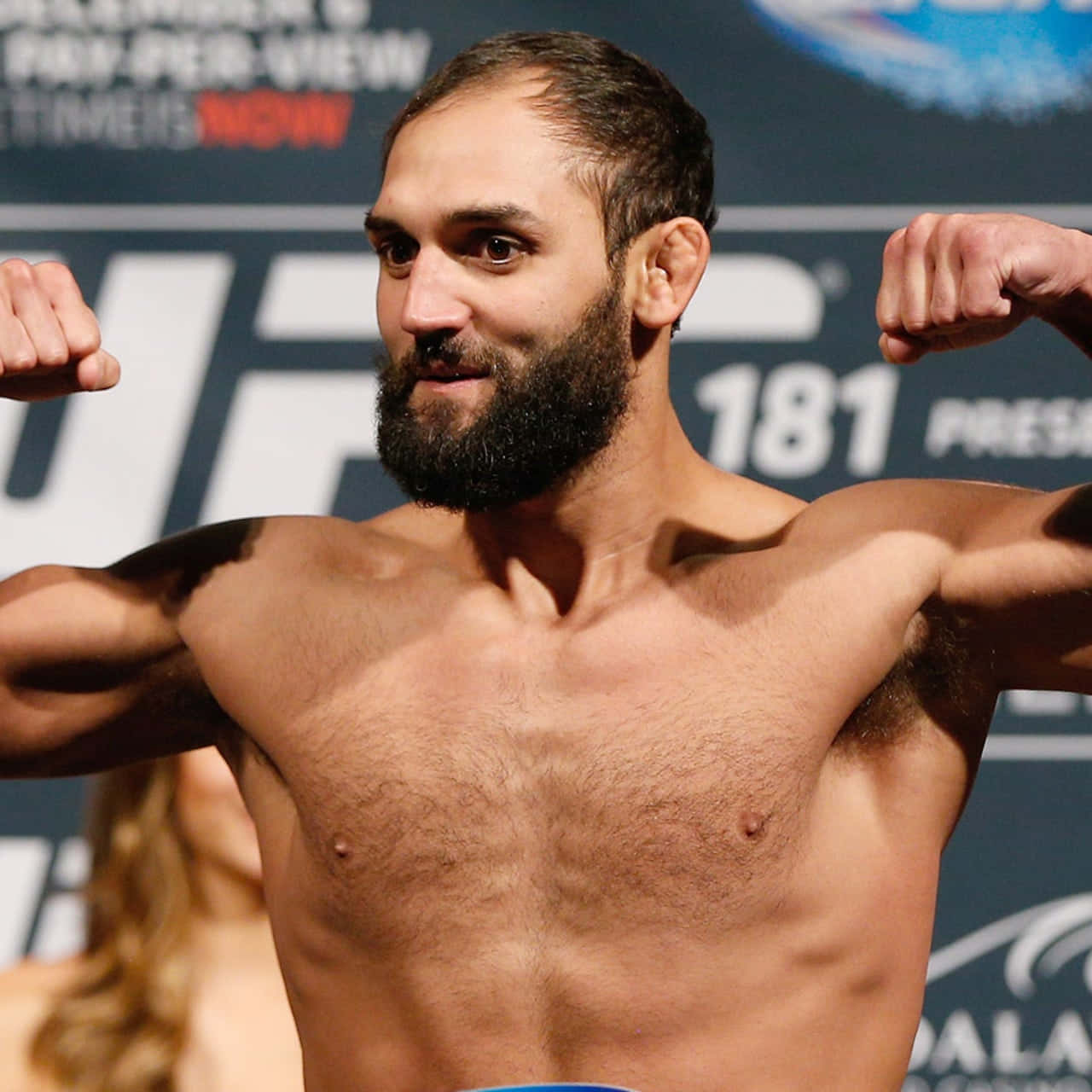 UFC 桌布 壁紙 — 一位肌肉發達、留鬍子的拳手在明亮的燈光下自信地彎曲手臂，參加 UFC 稱重活動。