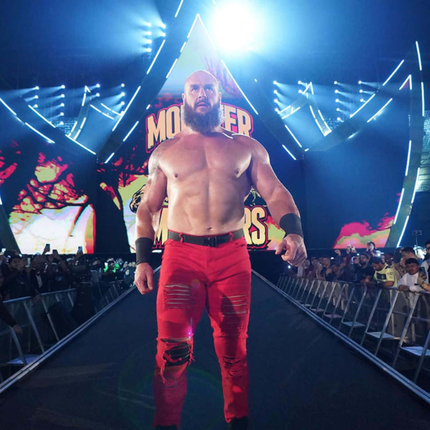 WWE 桌布 壁紙 — 一位肌肉發達、留鬍子的摔角手穿著破爛的紅色褲子，在明亮的舞台燈光下沿著明亮的競技場坡道走向咆哮的粉絲。