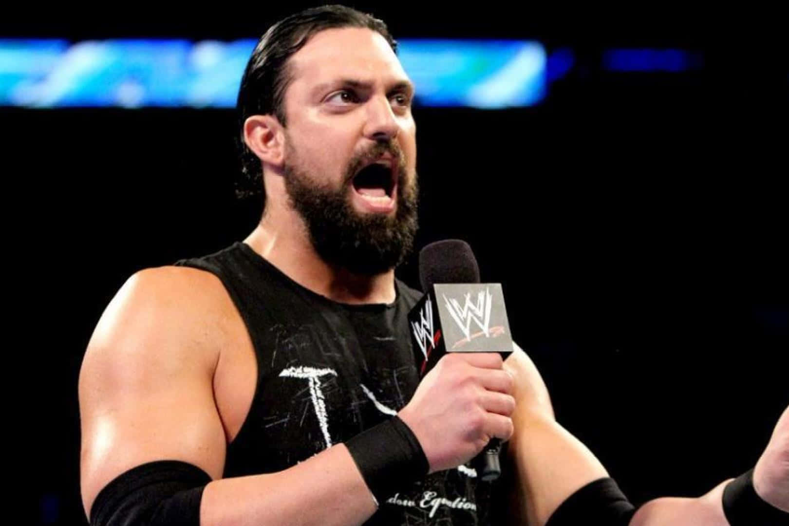 WWE 桌布 壁紙 — 一位肌肉發達、留鬍子的摔角手穿著黑色背心，在明亮的場館燈光下對著WWE麥克風大喊。