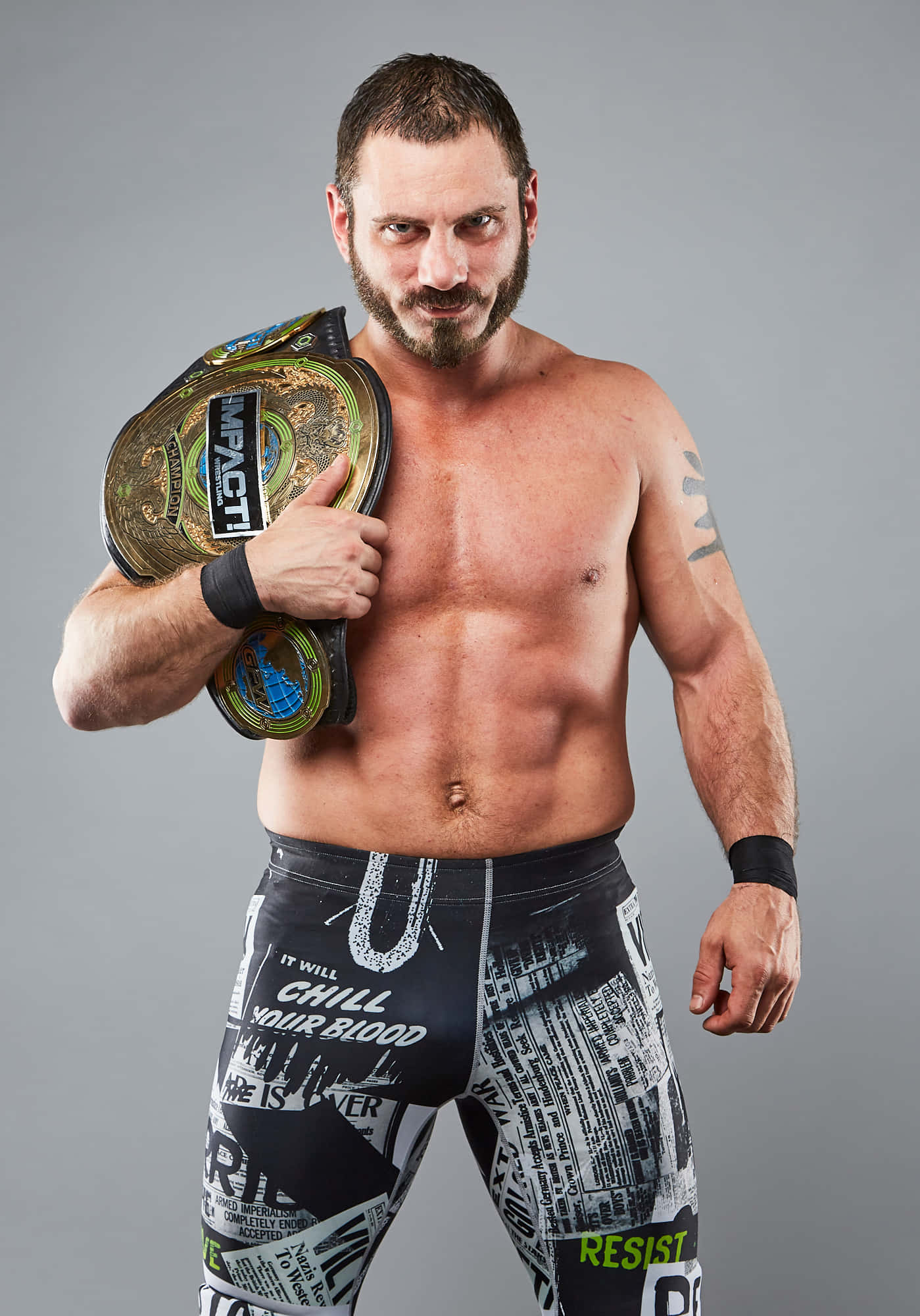 Champion 桌布 壁紙 — 一位肌肉發達、留鬍子的摔角手穿著圖案短褲，在純灰色工作室背景前舉起一條金綠色的 Impact 冠軍腰帶。