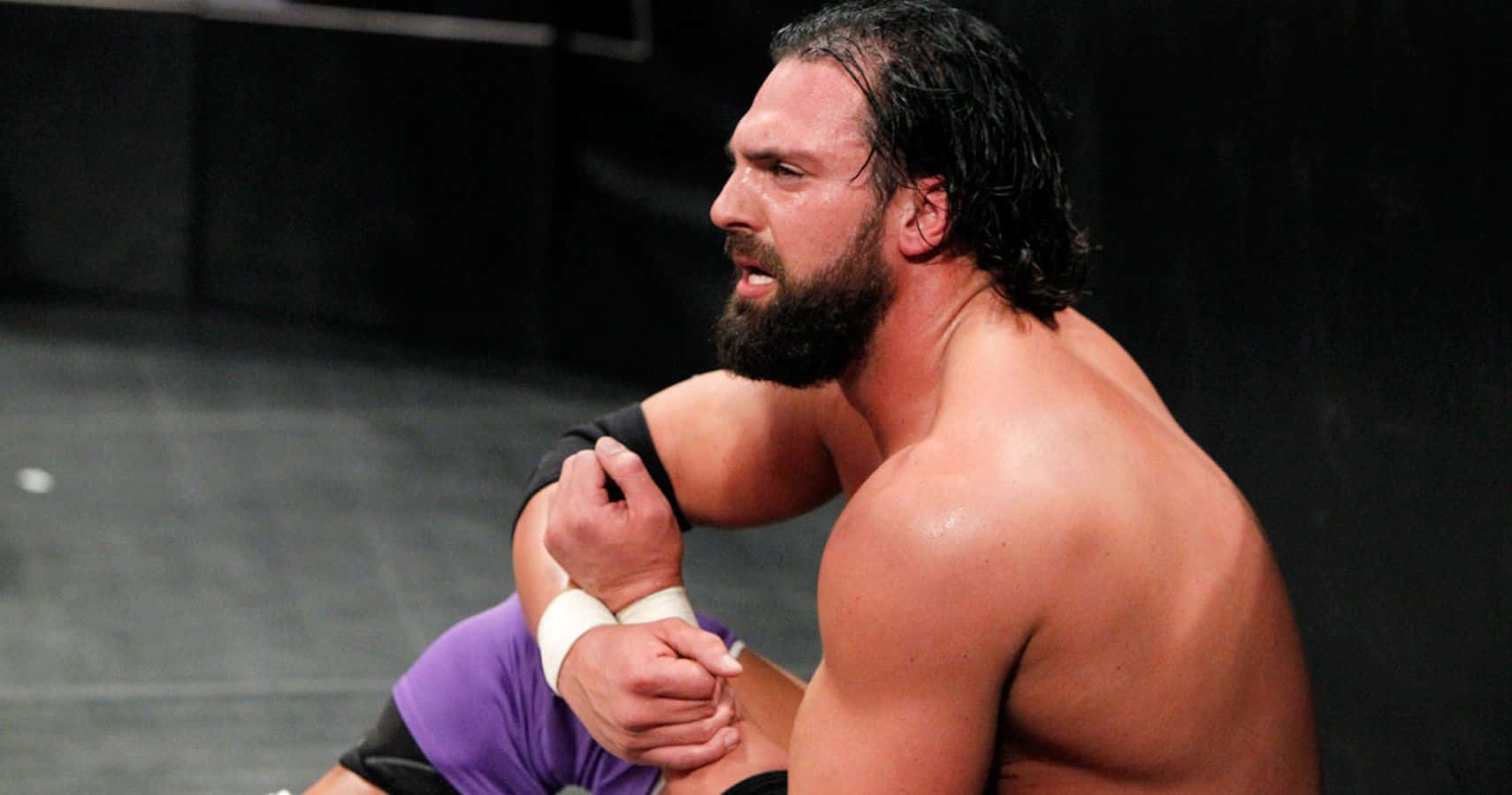 WWE 桌布 壁紙 — 一位肌肉發達、留鬍子的摔角手穿著紫色緊身褲坐在墊子上，專注地抓住自己的手臂。