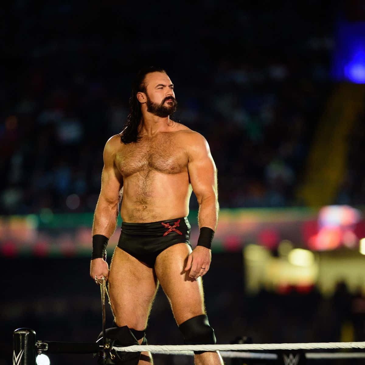 WWE 桌布 壁紙 — 一位肌肉發達、留鬍子的摔角手站在擂台上，在場館燈光下拿著一條鐵鏈。