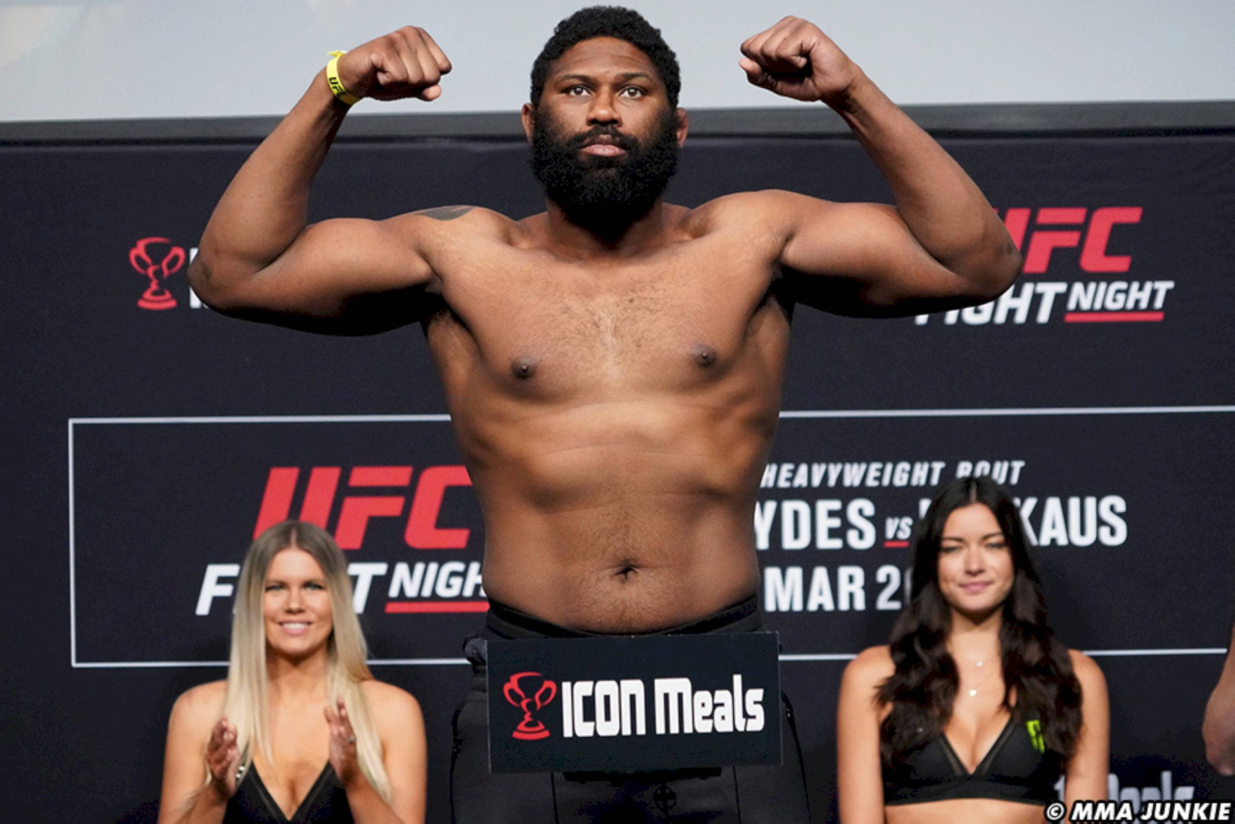 MMA 桌布 壁紙 — 一位肌肉發達、留鬍子的格鬥家在UFC稱重時赤裸上身展示肌肉，旁邊有兩位微笑的比基尼女郎。