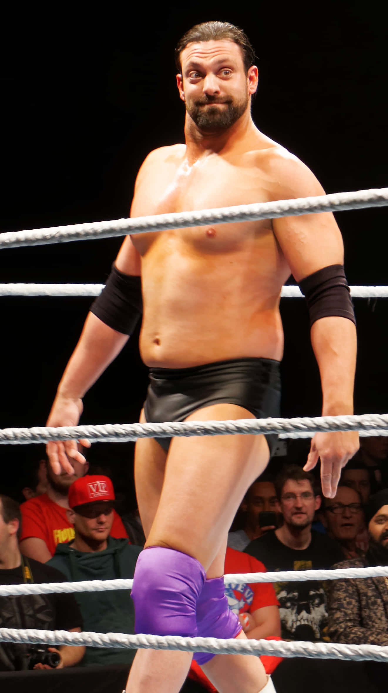 WWE 桌布 壁紙 — 一位肌肉發達、留鬍鬚、戴紫色護膝的摔角手，自信地站在有繩索和觀眾背景的摔角台上。
