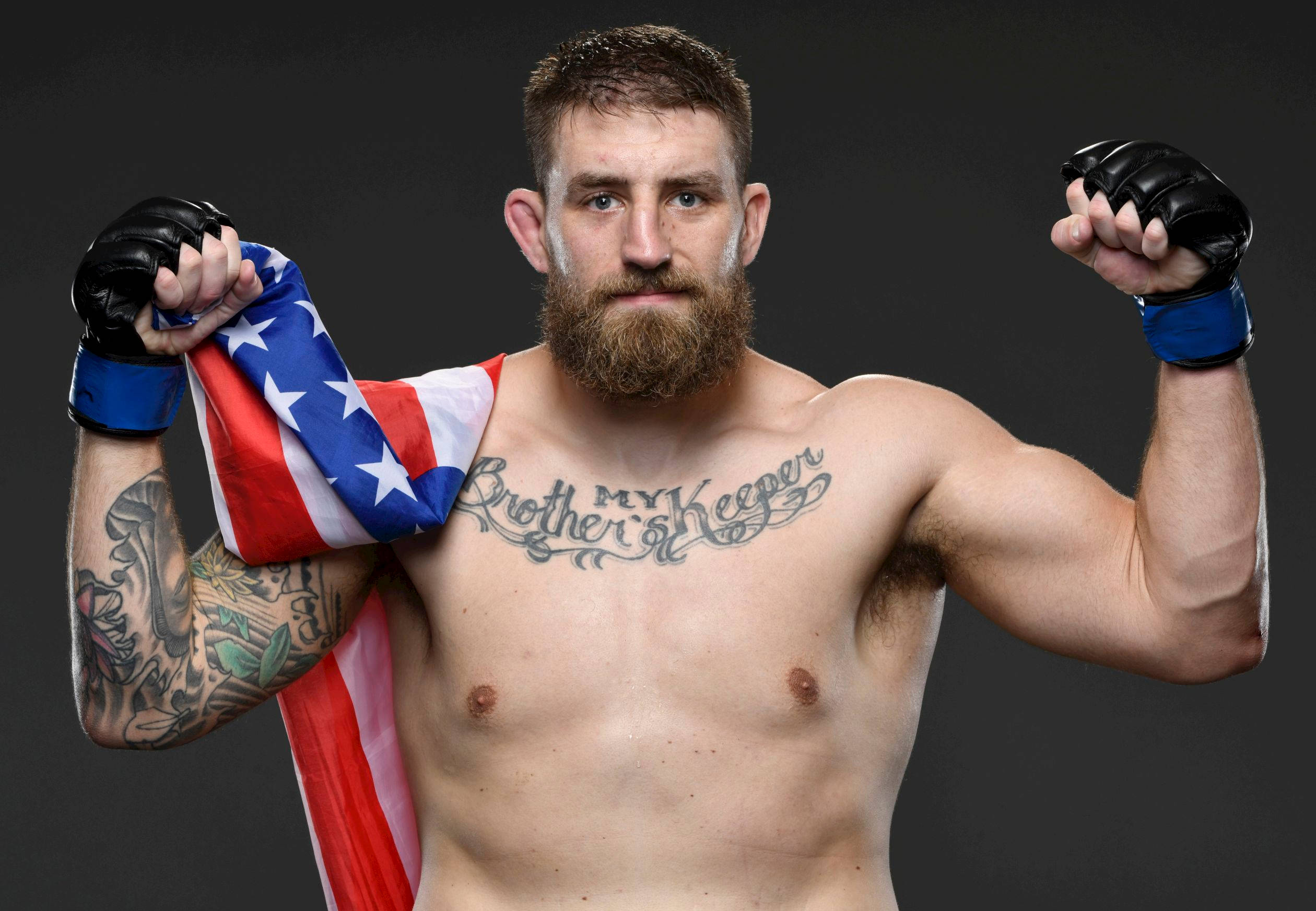 UFC 桌布 壁紙 — 一位肌肉發達、留鬍鬚、有刺青的戰士，在黑暗的攝影棚背景下，自豪地將美國國旗扛在肩上擺姿勢。