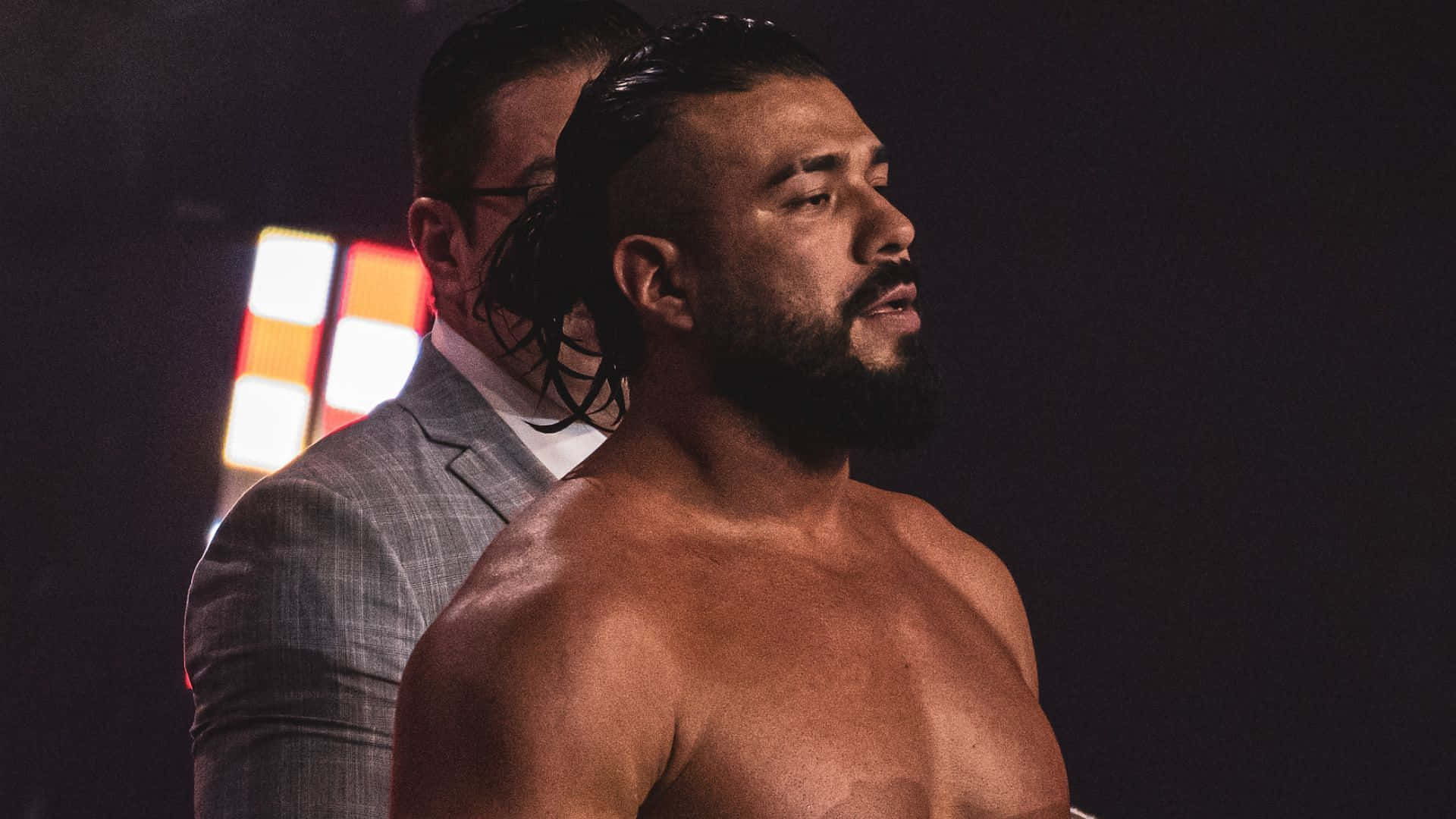 Aew 桌布 壁紙 — 一位肌肉發達、留鬍鬚的摔角手，濕髮赤裸上身站在戲劇性的舞台燈光下，身後有一位穿西裝的男子。