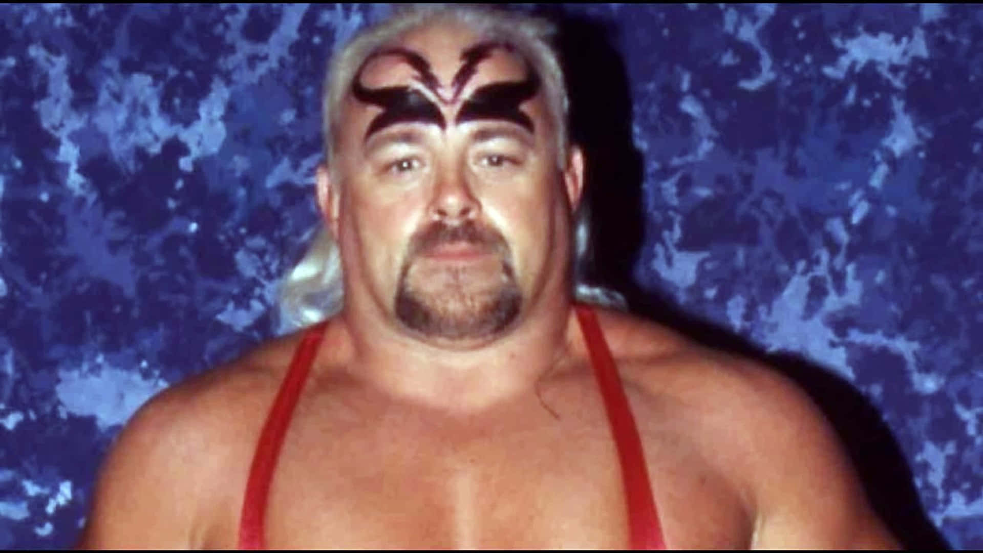WWE 桌布 壁紙 — 一位肌肉發達、白髮黑臉的摔角手，穿著紅色緊身衣，在帶有紋理的藍色背景前擺姿勢。