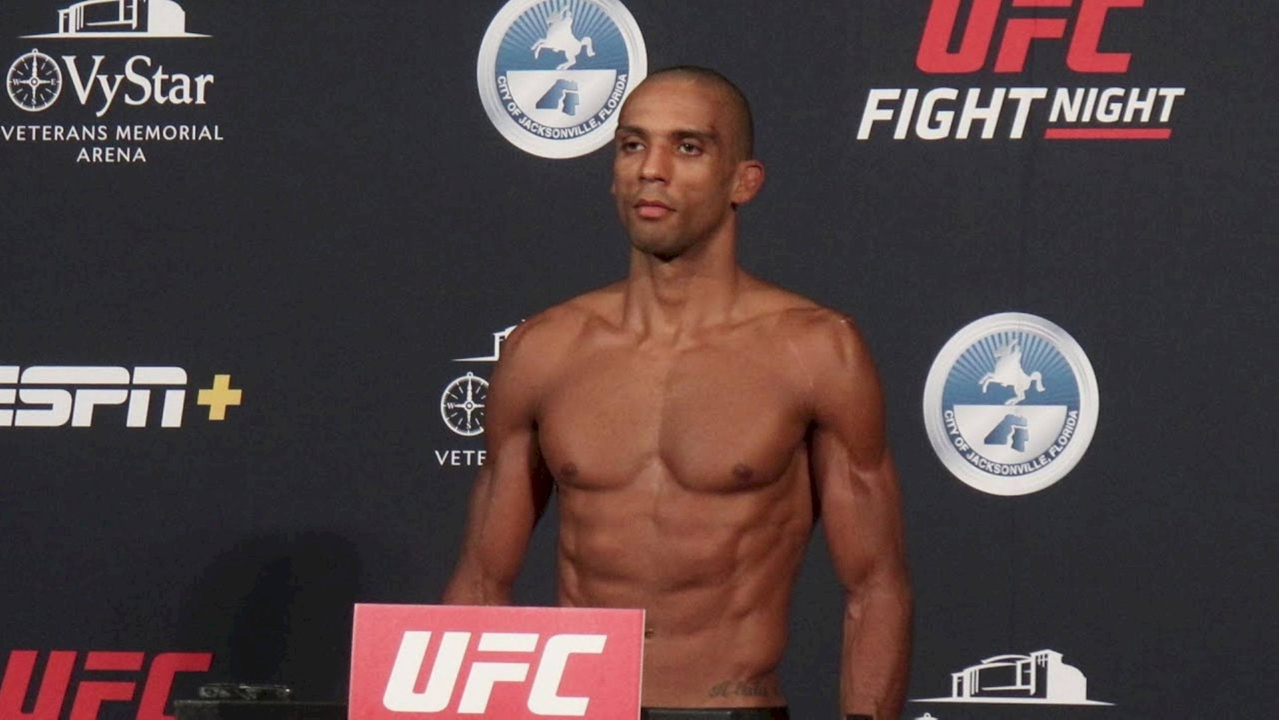 UFC 桌布 壁紙 — 一位肌肉發達、赤膊的拳手自信地站在有品牌背景標誌的UFC稱重活動中。