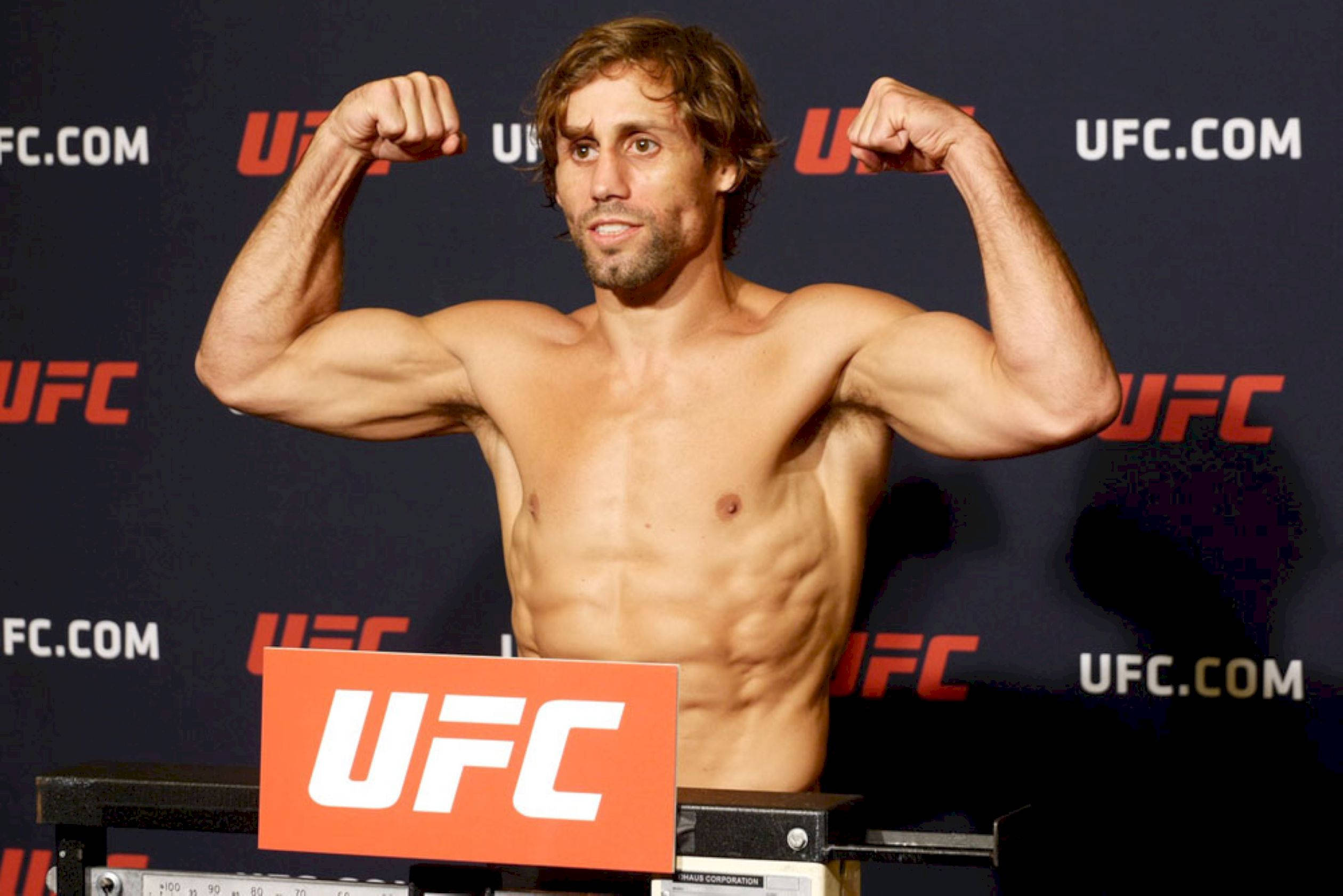 MMA 桌布 壁紙 — 一位肌肉發達、赤裸上身的拳手在UFC稱重儀式上，雙臂彎曲，背景有品牌秤台。