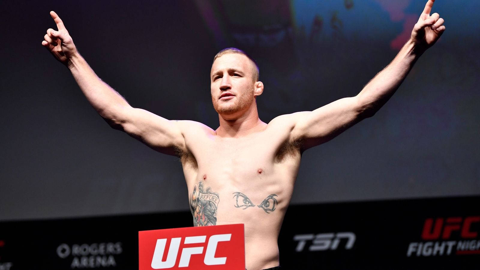 UFC 桌布 壁紙 — 一位肌肉發達、赤裸上身的拳手在明亮的燈光下，在UFC擂台上勝利地高舉雙臂，手指向上指著。