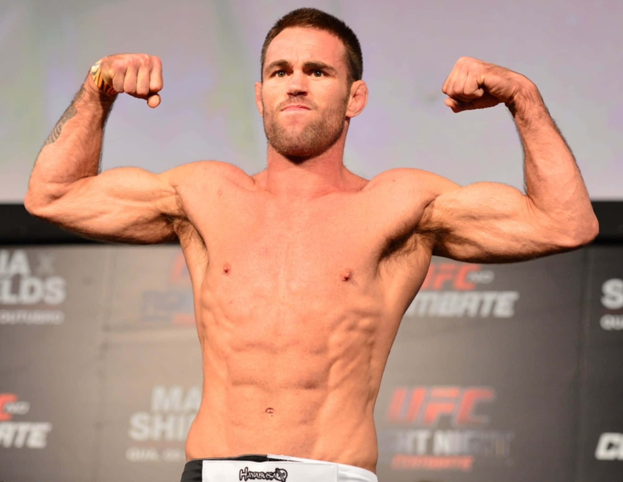 Athletics 桌布 壁紙 — 一位肌肉發達、赤裸上身的拳手在明亮的舞台燈光下，於 UFC 稱重活動中彎曲雙臂。