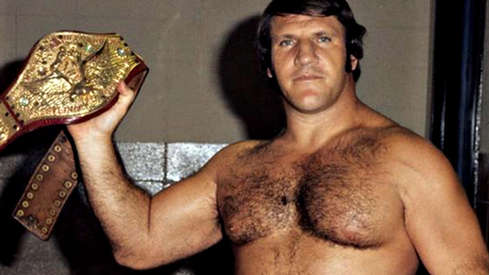 Champion 桌布 壁紙 — 一位肌肉發達、赤裸上身的摔角手在昏暗的房間裡手持金色冠軍腰帶，專注地凝視著鏡頭。