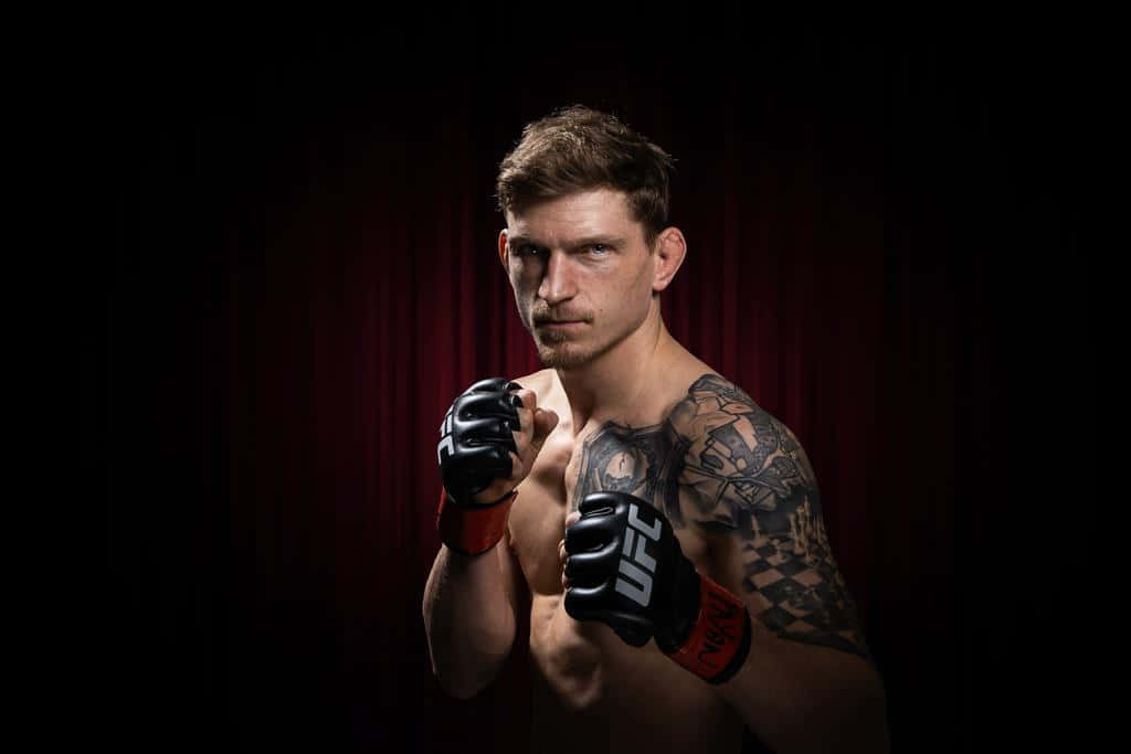 UFC 桌布 壁紙 — 一位肌肉發達、身上有刺青的拳手，戴著UFC手套，在深紅色窗簾背景下凝視著。