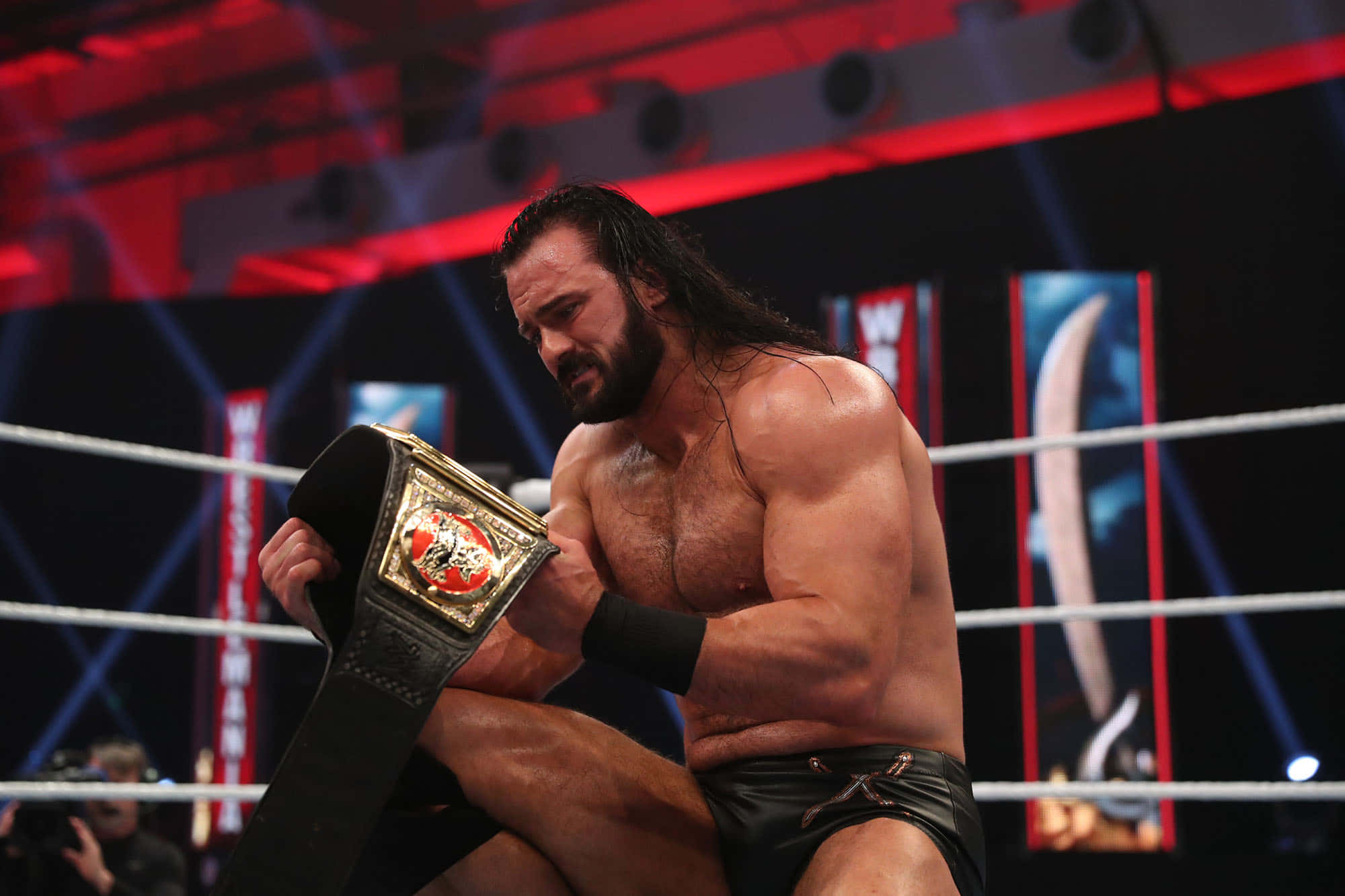 WWE 桌布 壁紙 — 一位肌肉發達、長黑髮的摔角手坐在擂台上，在戲劇性的紅色舞台燈光下手持冠軍腰帶。