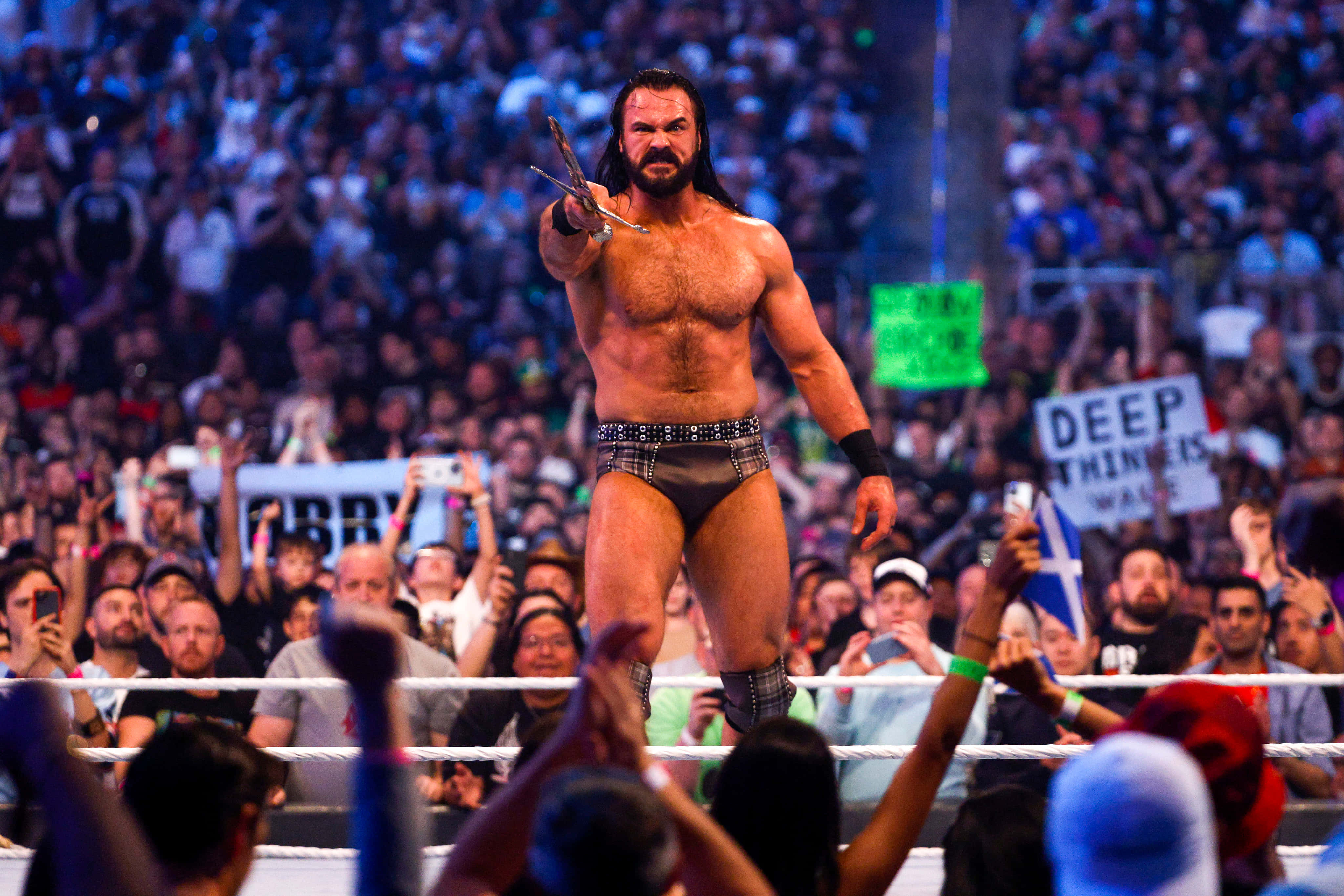 WWE 桌布 壁紙 — 一位肌肉發達、長黑髮的摔角手站在擂台上，在明亮的場館燈光下，拿著一把剪刀指向人群。