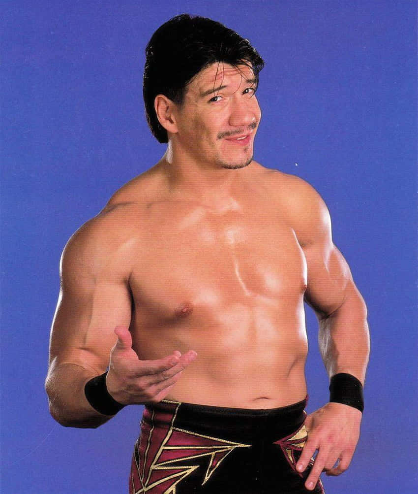 Wrestling 桌布 壁紙 — 一位肌肉發達、黑髮留鬍的摔角手，自信地擺姿勢，背景是純藍色攝影棚，穿著黑紅圖案短褲。
